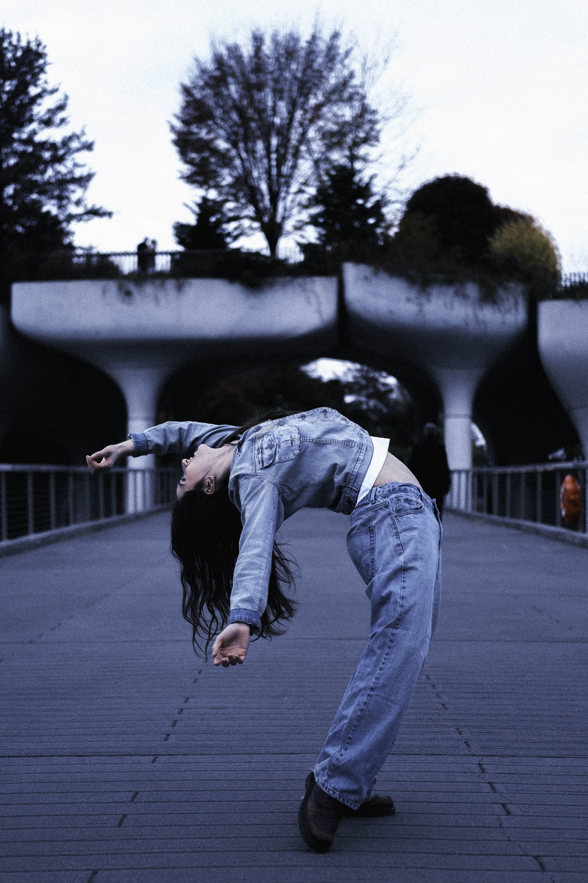 A young person with long dark hair wearing a denim jacket and jeans, mid-motion, bending backward on a bridge with a railing. In the background, there are trees and a bridge with arches over a waterway, with an overcast sky.