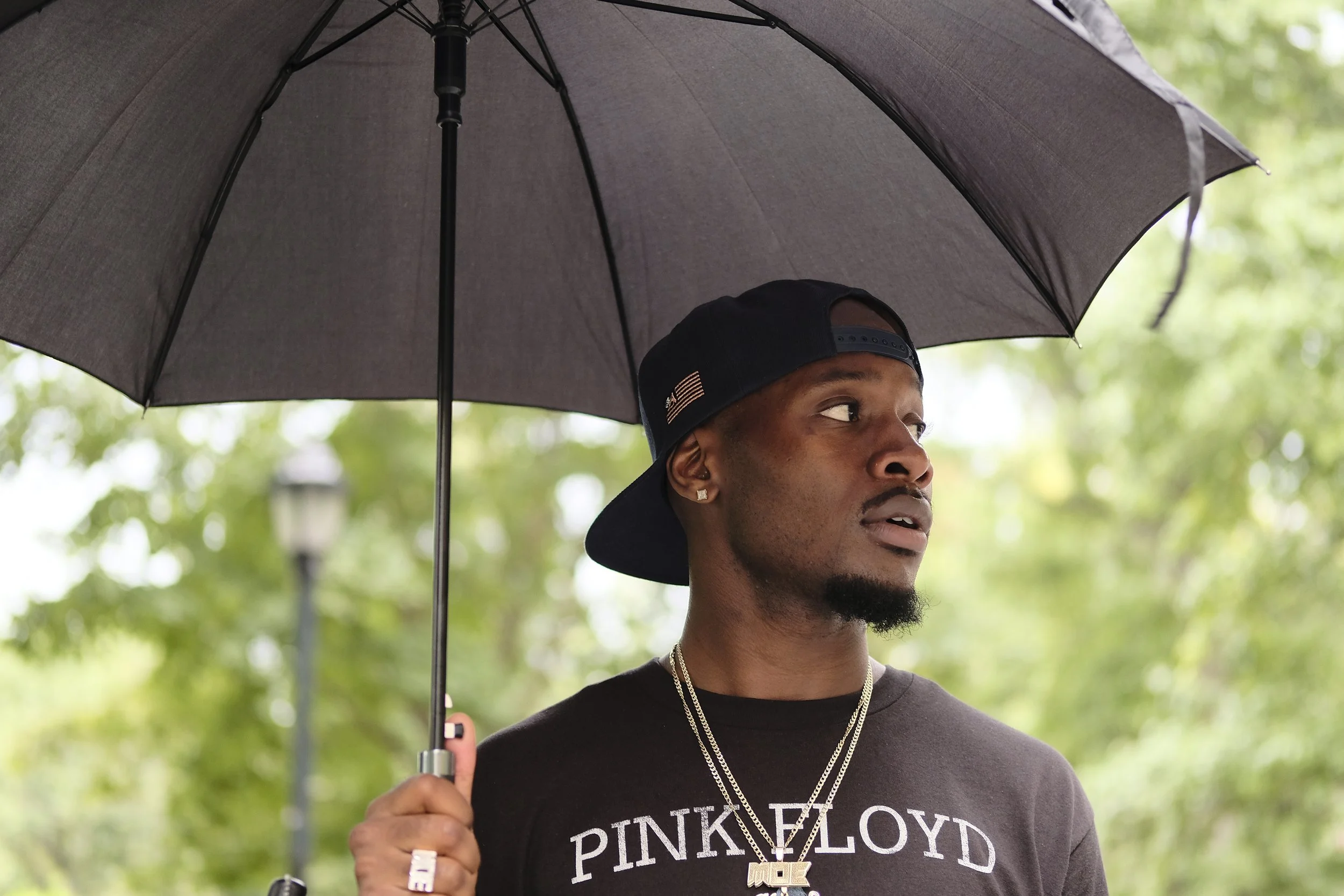 A man with dark skin, wearing a black Pink Floyd t-shirt, a black cap worn backwards, gold chains, and earrings, is holding a black umbrella outdoors. He is looking to the right, and there are green trees in the background.