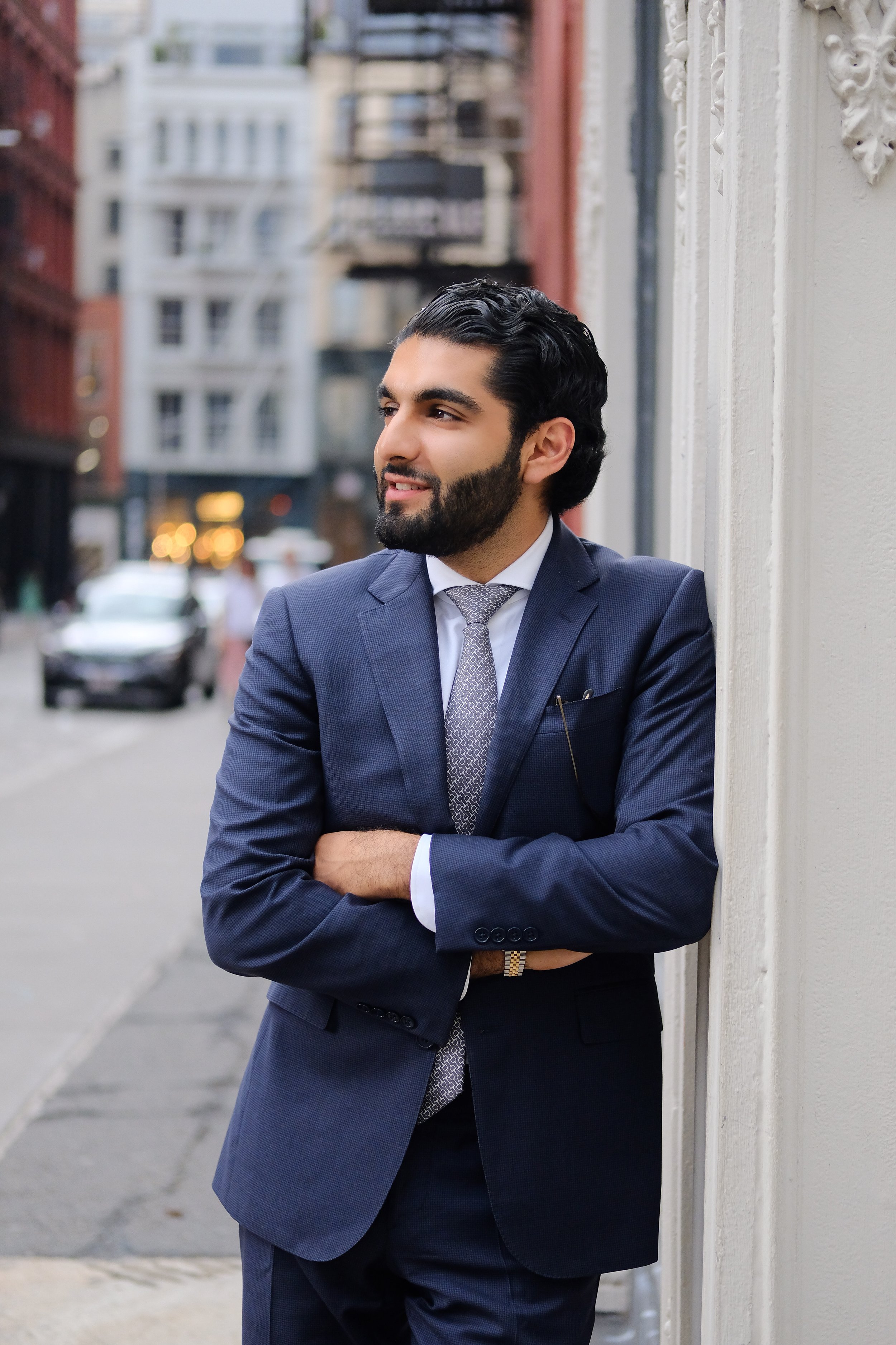 A man in a navy suit with a tie is standing outdoors against a white wall, with his arms crossed, looking to his left, with buildings and blurred cars in the background.