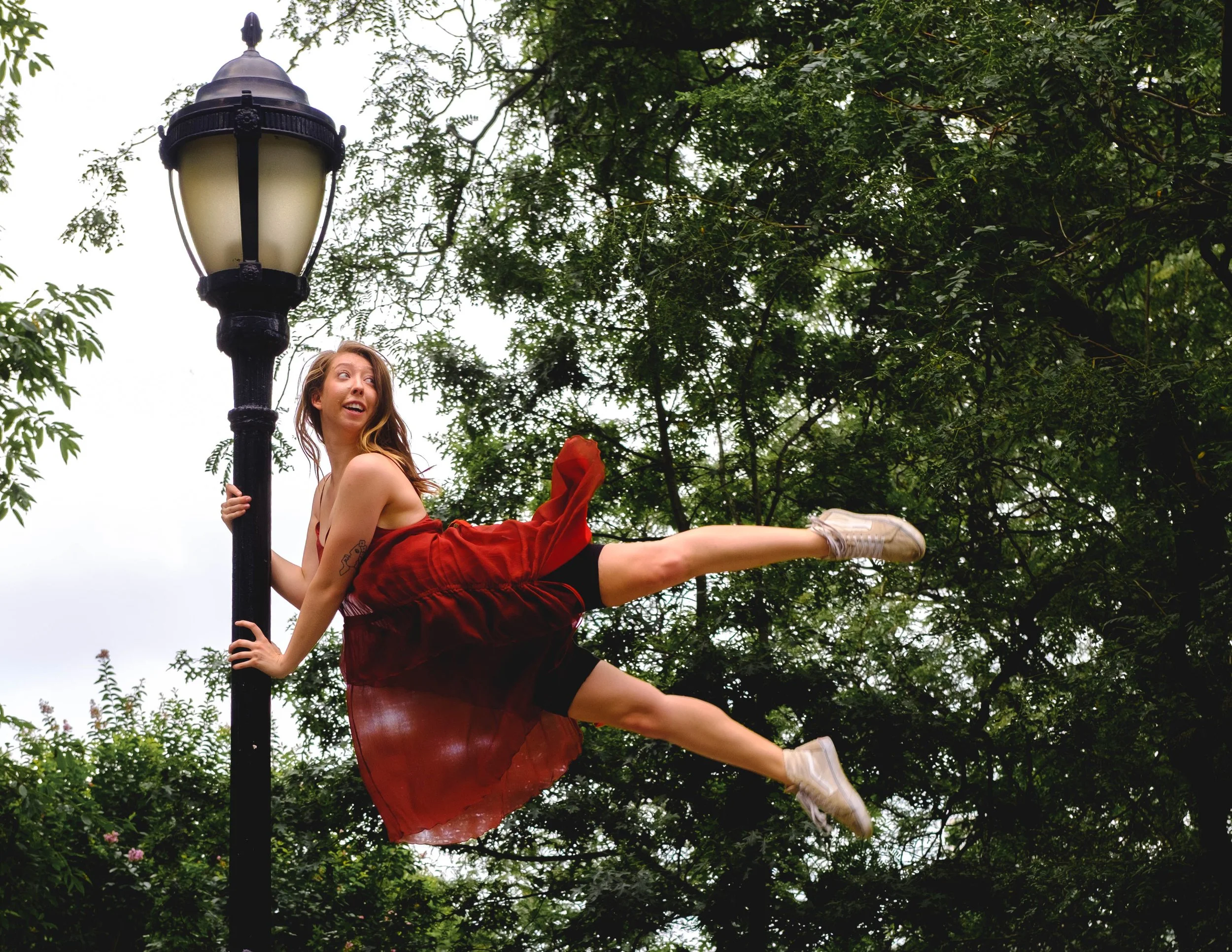 A young woman sitting on a swing attached to a streetlamp post, wearing a red dress and sneakers, with trees and green foliage surrounding her.