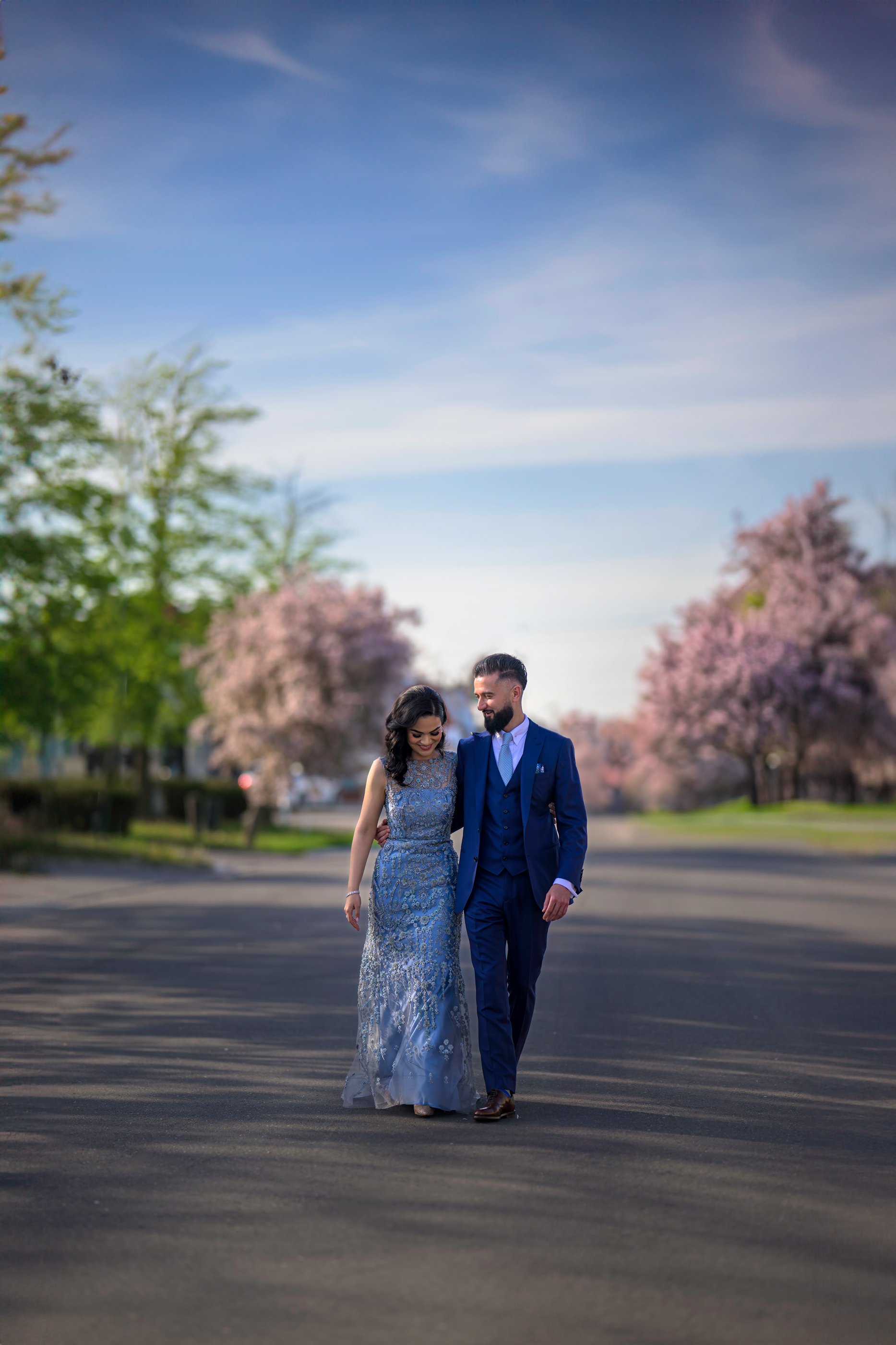 A couple walking arm in arm on a paved path through a park with pink flowering trees, blue sky, and some green trees in the background.