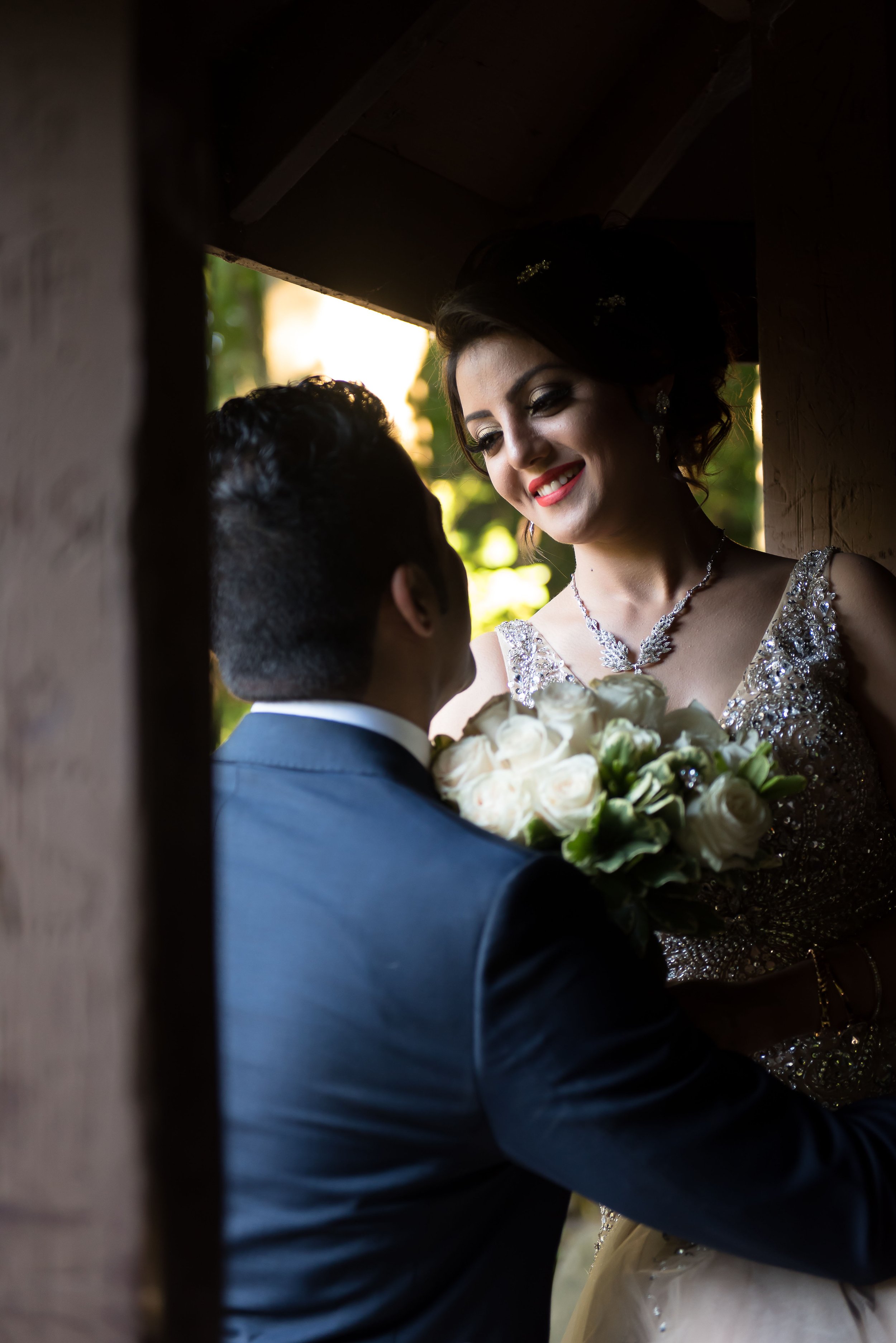 Bride and groom on their wedding day, smiling at each other. The bride holds a bouquet of white roses, wearing a sparkling dress and jewelry. The groom wears a dark tuxedo.