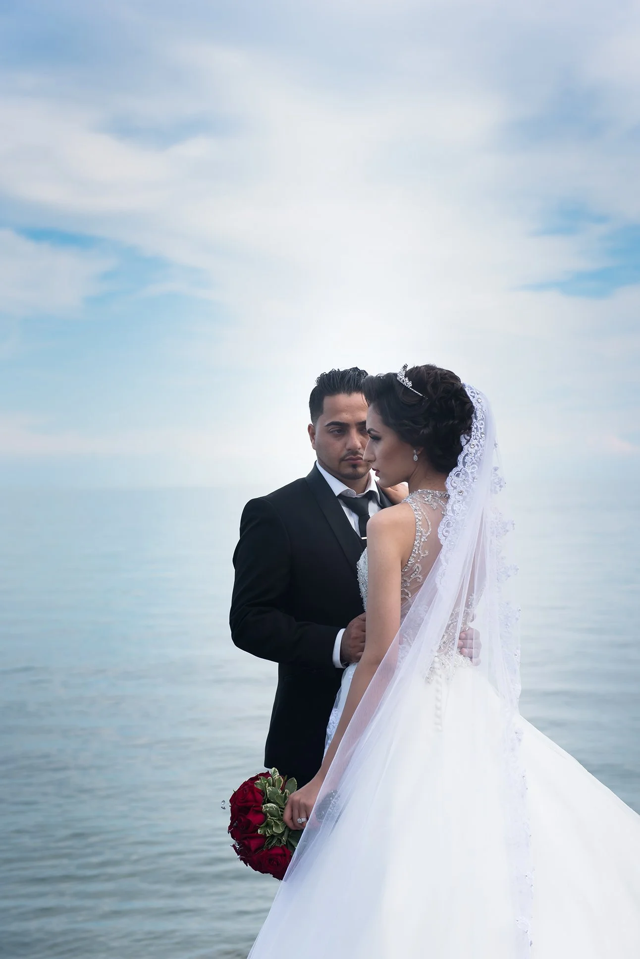 Wedding couple standing by water, the bride in a white gown with a veil holding a bouquet of red roses, the groom in a black suit, against a cloudy sky and calm water background.