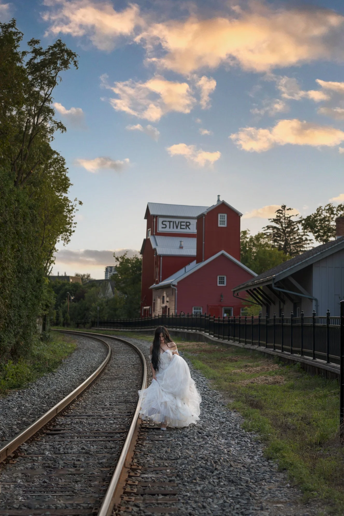 A woman in a white bridesmaid dress standing on a curved railway track with a red grain elevator and a blue sky with clouds in the background.