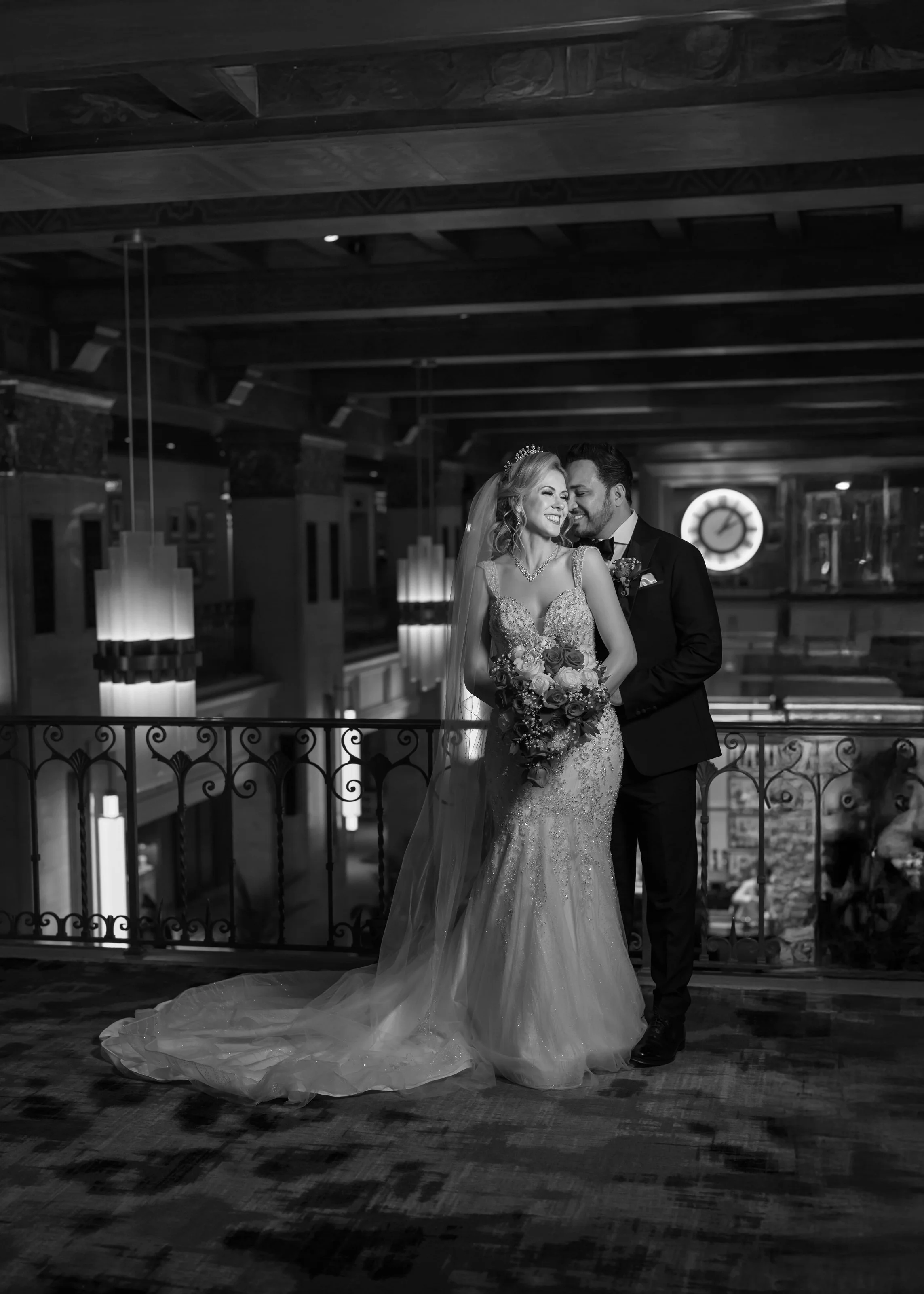 Black and white wedding photo of a bride and groom smiling and embracing on a balcony inside a decorated venue, with a large clock in the background.
