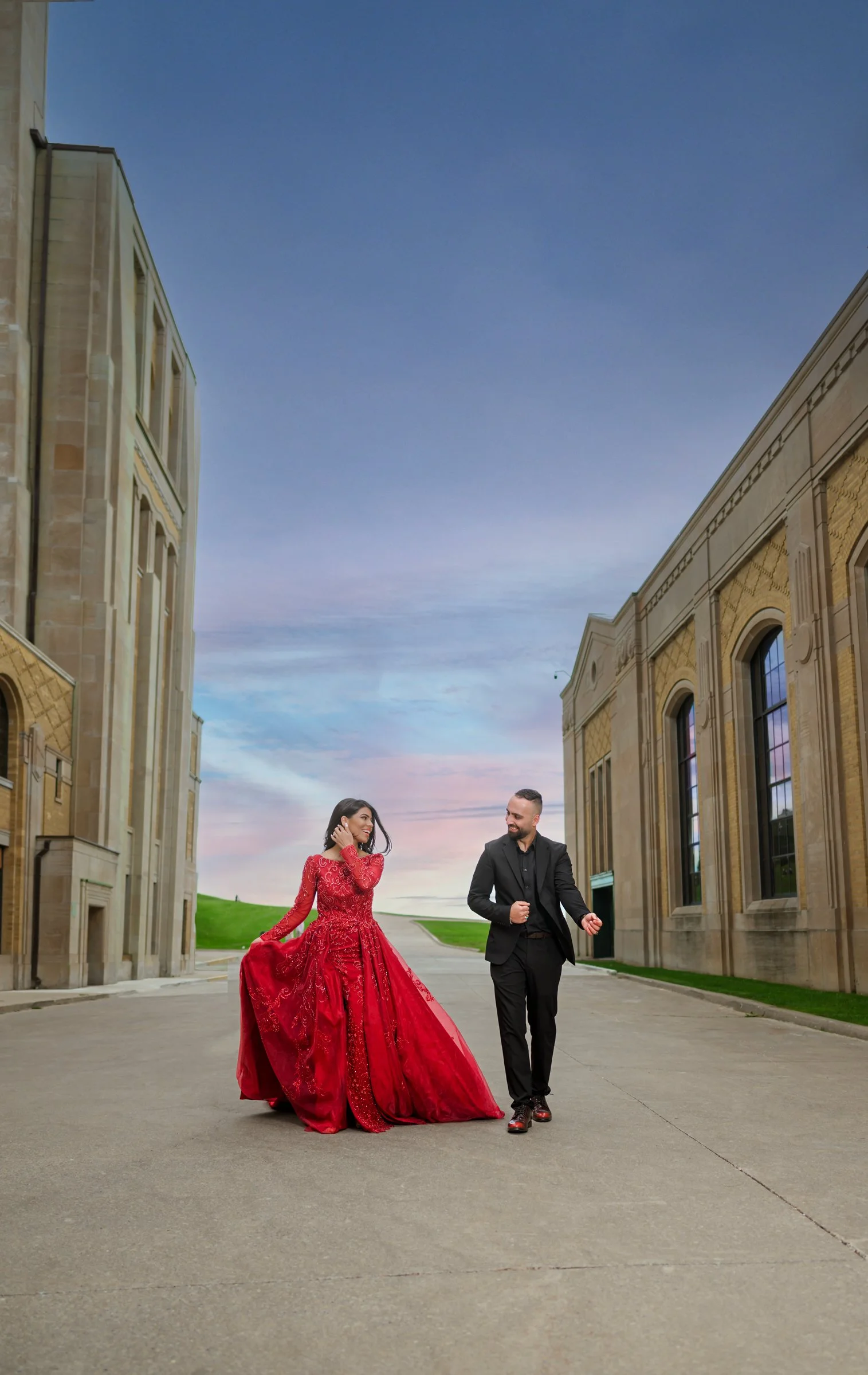 A woman in a red ball gown and a man in a black suit walking together on a paved street between historic buildings, with a colorful sky in the background.
