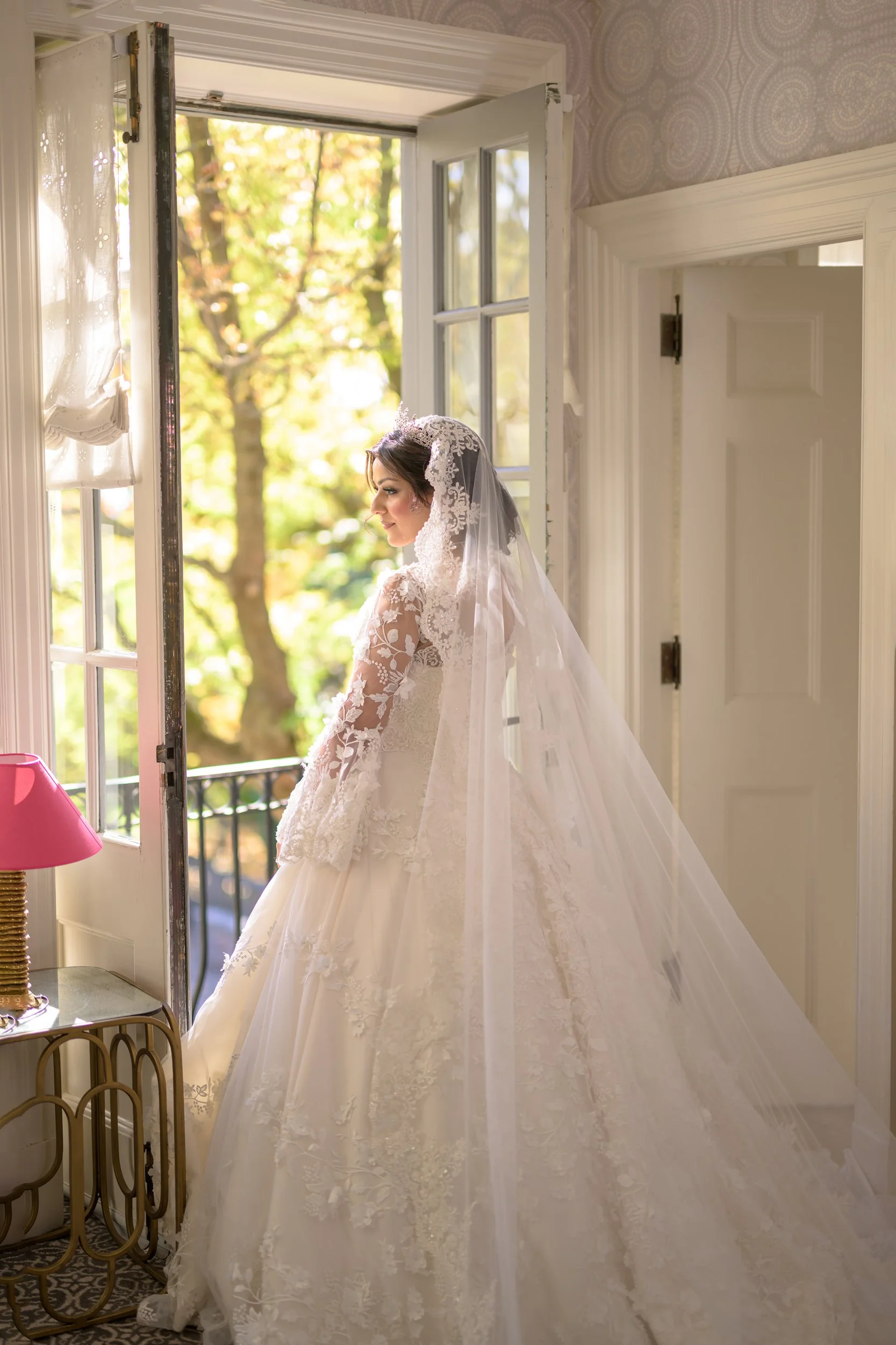 Bride standing by open window with sunlight streaming in, wearing a lace wedding dress and veil, in a cozy room with patterned wallpaper.