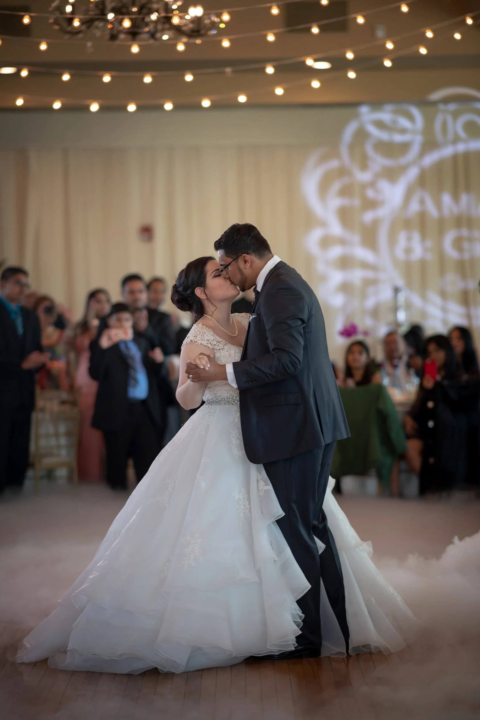A bride and groom share their first dance at their wedding reception, surrounded by seated guests in a decorated venue illuminated by string lights.