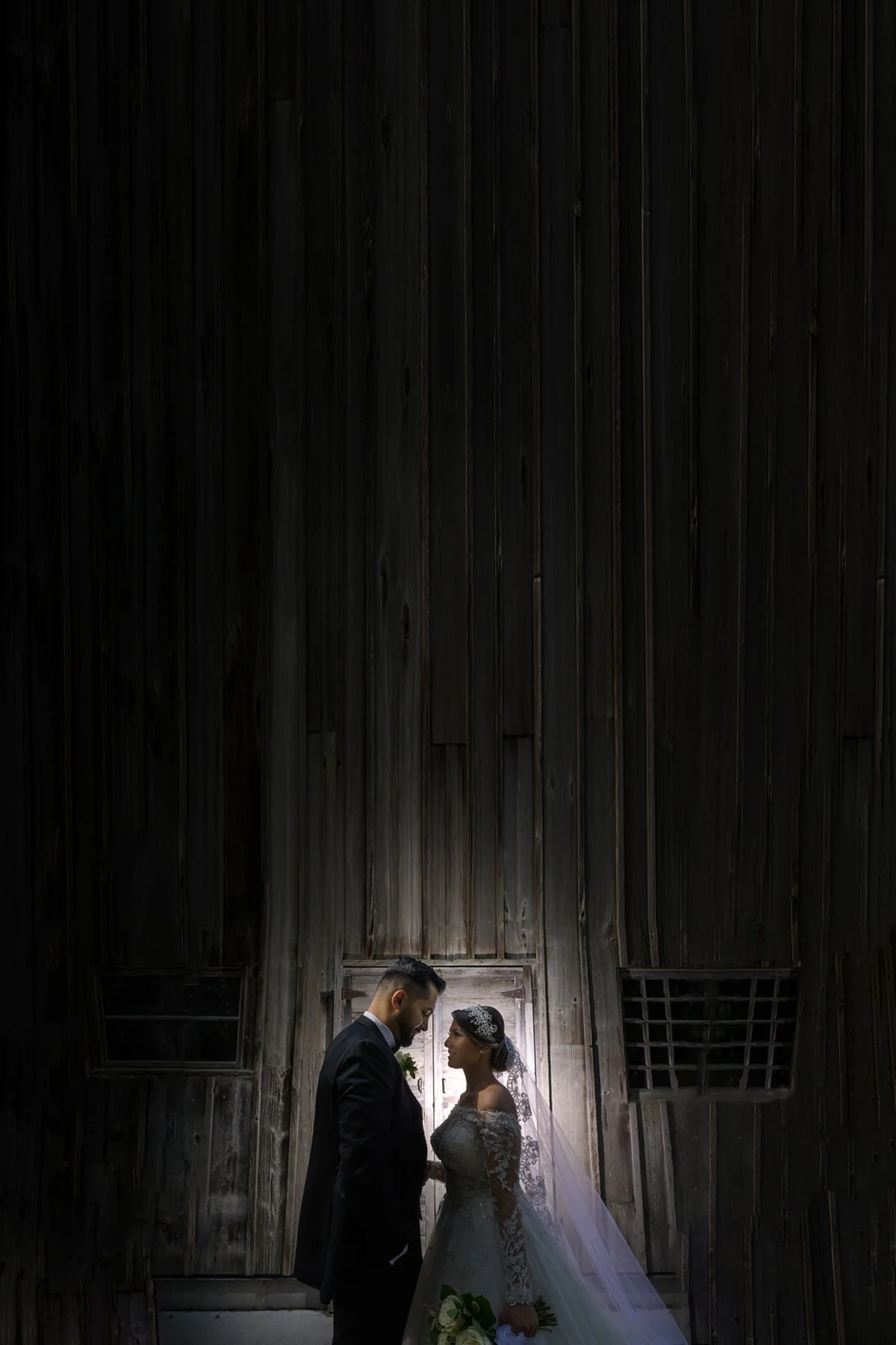 A bride and groom stand close together in front of a rustic wooden wall, illuminated by a dramatic light source from behind, creating silhouettes.