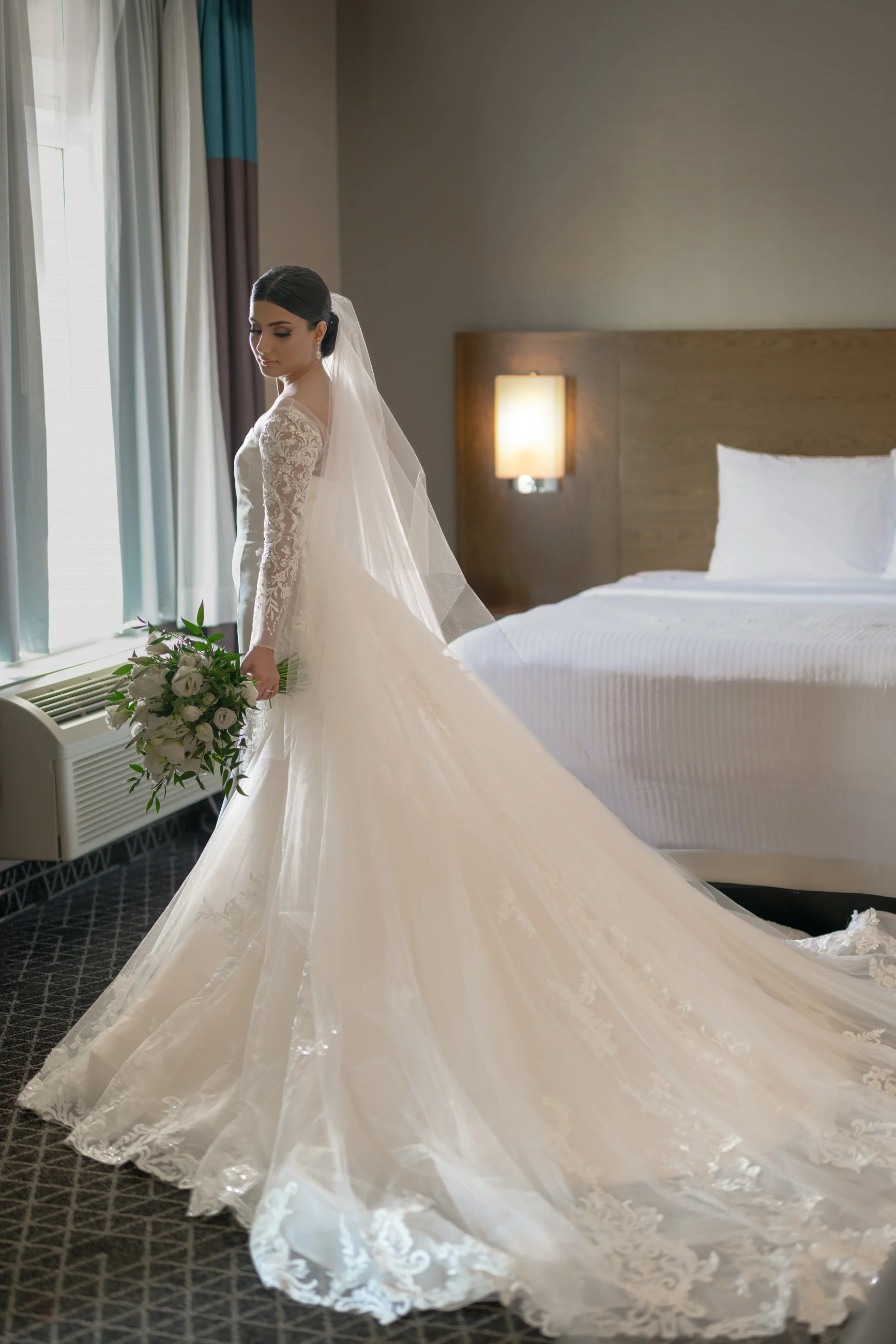 A bride in a white lace wedding dress with a long train and veil, holding a bouquet of white flowers, standing in a hotel room near a window with curtains.