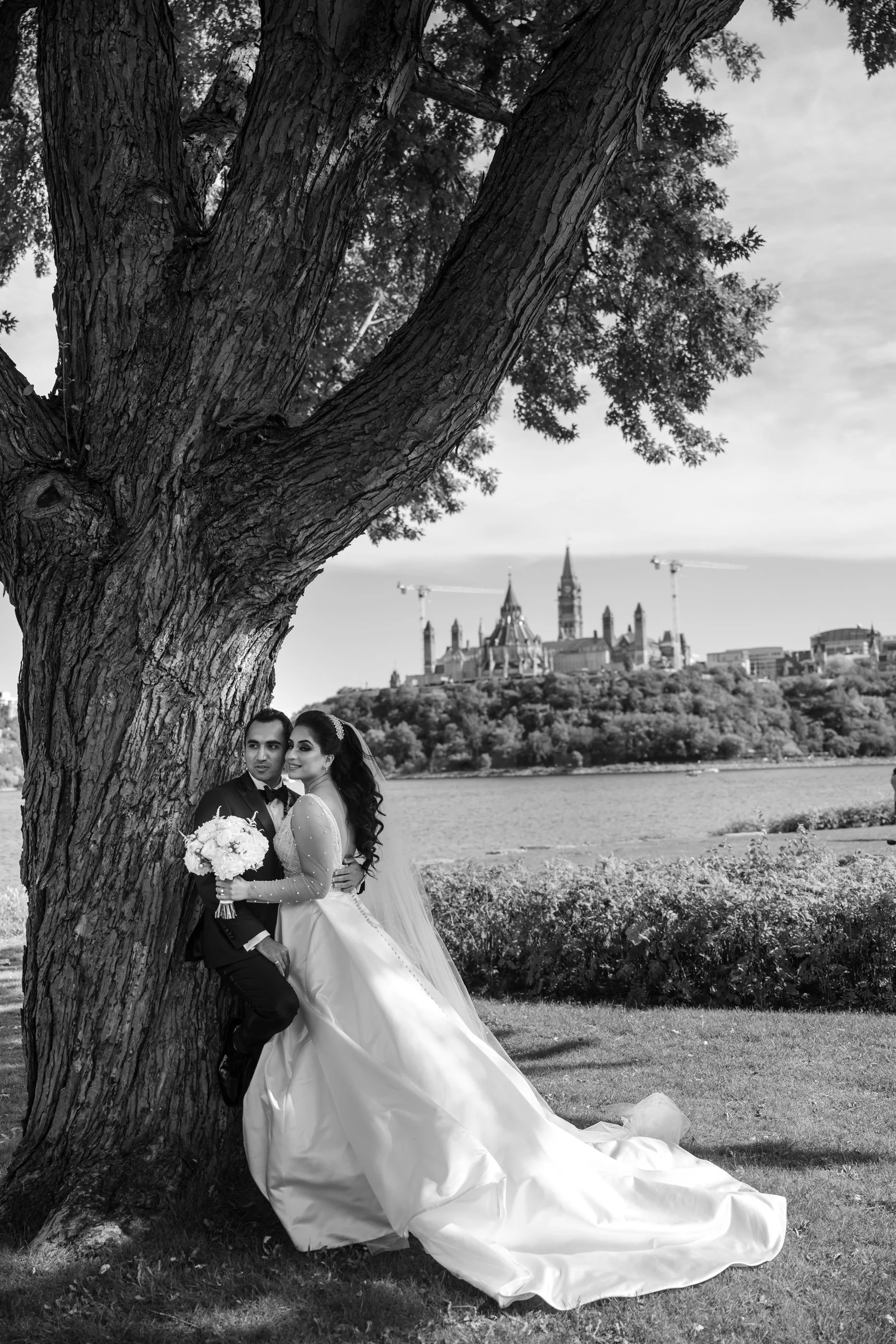 Black and white photo of a newlywed couple sitting under a large tree, with a river and castle in the background.