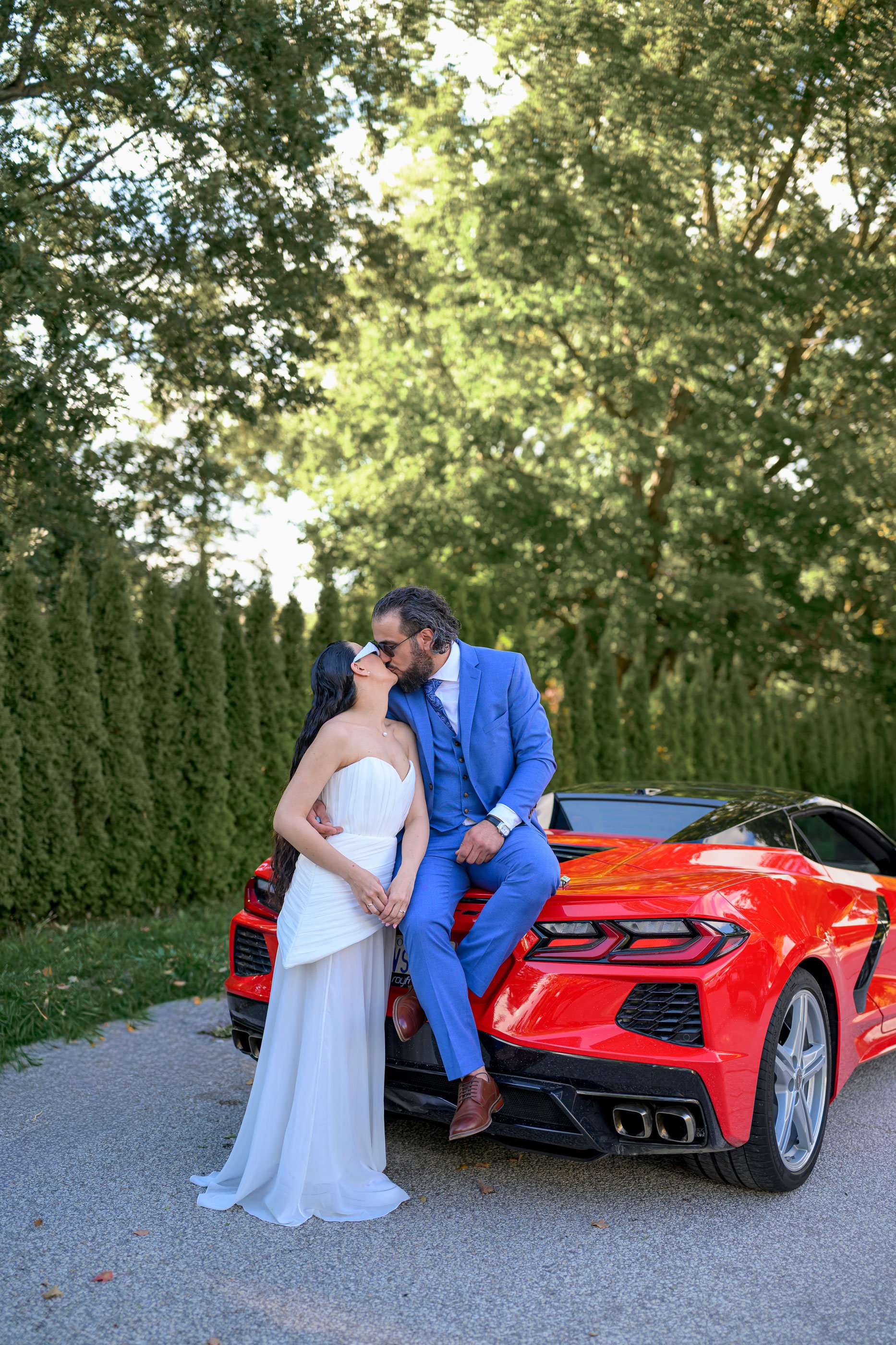 A couple in wedding attire sharing a kiss in front of a red sports car.