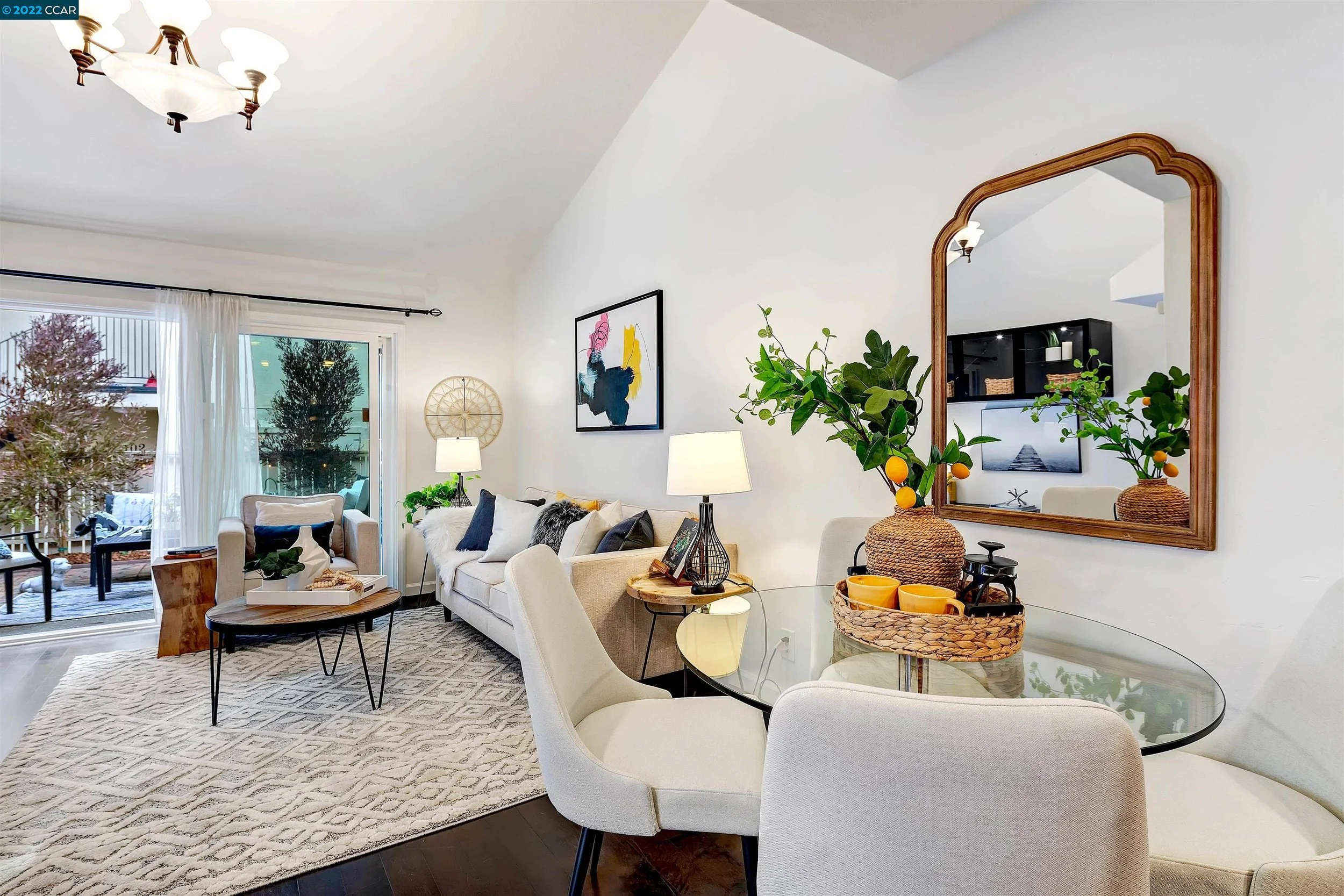 Living room with white sofa, armchair, glass dining table, and sliding glass door leading to outdoor patio with trees.