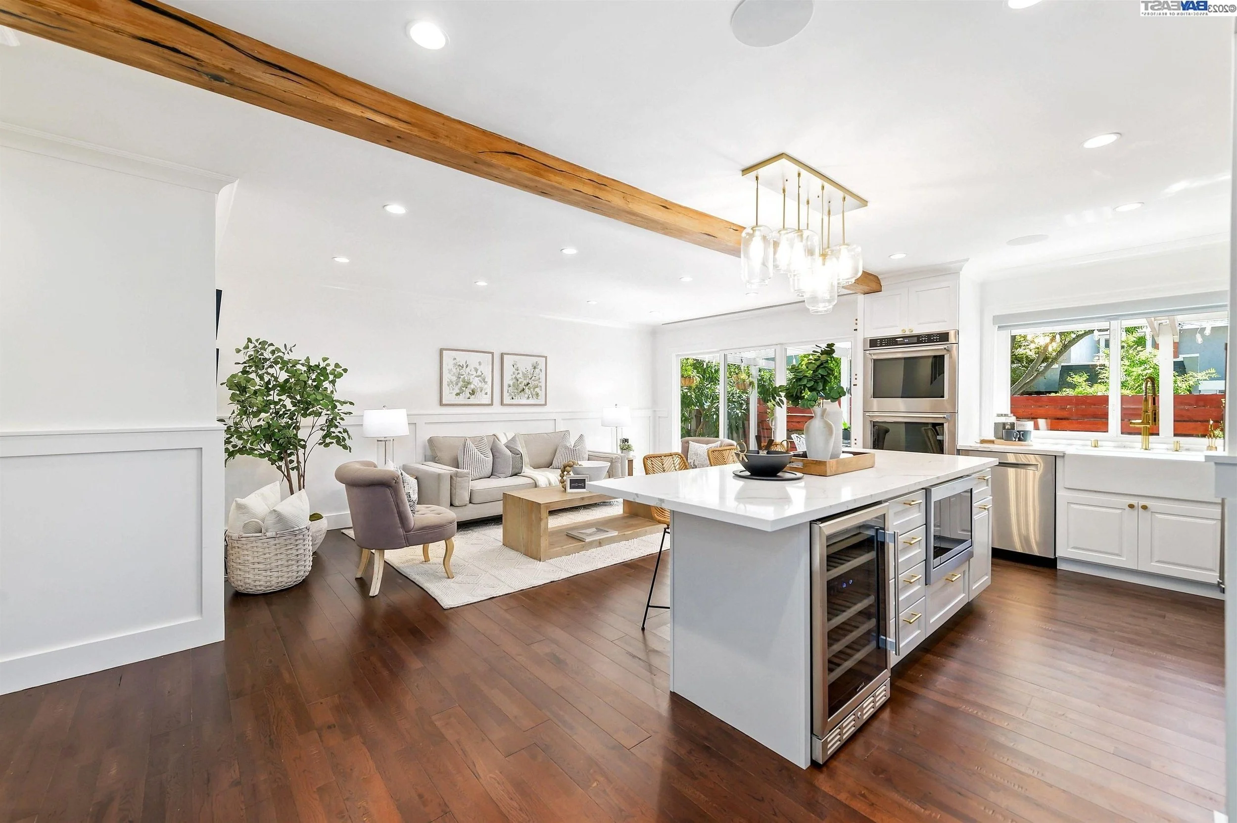 Open-concept kitchen and living room with white walls, wooden floors, and modern decor. The kitchen features a large white island with a plant in a white vase, and built-in appliances. The living area has a beige sofa, armchair, and a coffee table with framed botanical prints on the wall. Large windows let in natural light and reveal greenery outside.
