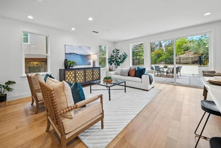 Living room with white walls, large windows, wooden floor, beige and white furniture, decorative pillows, a painting, plants, and a sliding glass door leading to a patio with outdoor furniture.