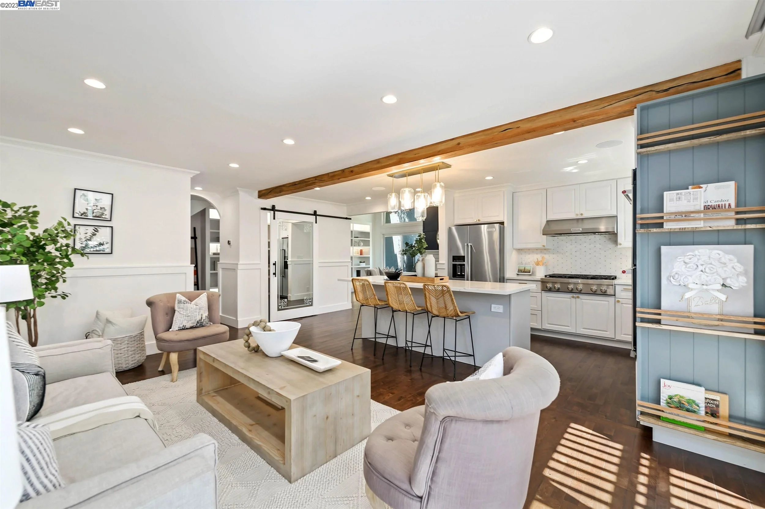 Open living room and kitchen area with white walls, hardwood floors, a beige sofa, armchairs, a wooden coffee table, and kitchen with white cabinets, stainless steel appliances, a white island with three wicker barstools, and a blue accent wall with shelves.