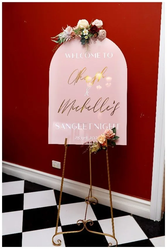 Wedding welcome sign on an easel with flowers, against a red wall, for a Sangeet Night event on September 28, 2023.