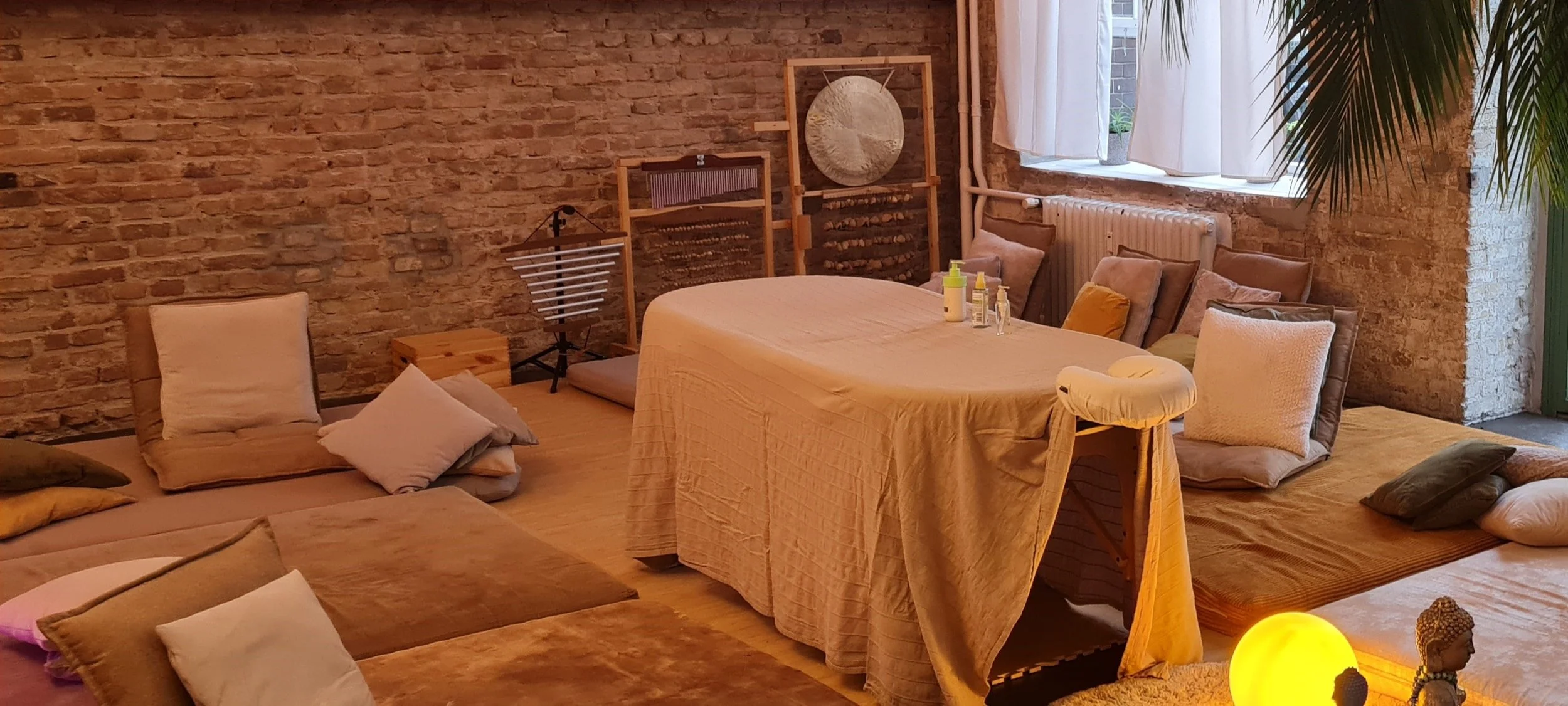 Innenraum mit einem runden Tisch, der mit einem hellen Tuch bedeckt ist, umgeben von Bodenkissen und Polstern. Im Hintergrund eine Ziegelsteinwand, Fenster mit weißen Vorhängen, Pflanzen und Dekorationen.