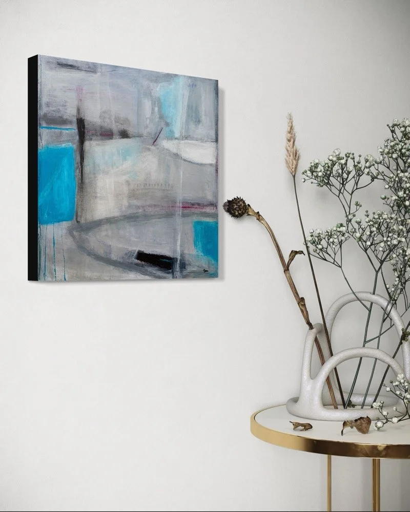 Gera Dunachie’s abstract painting "Held Back" shown in a deep black frame on a plain white wall. To the right, a sculptural, loop-shaped ceramic vase sits on a gold-rimmed side table, holding delicate dried stems. Minimalistic decor.
