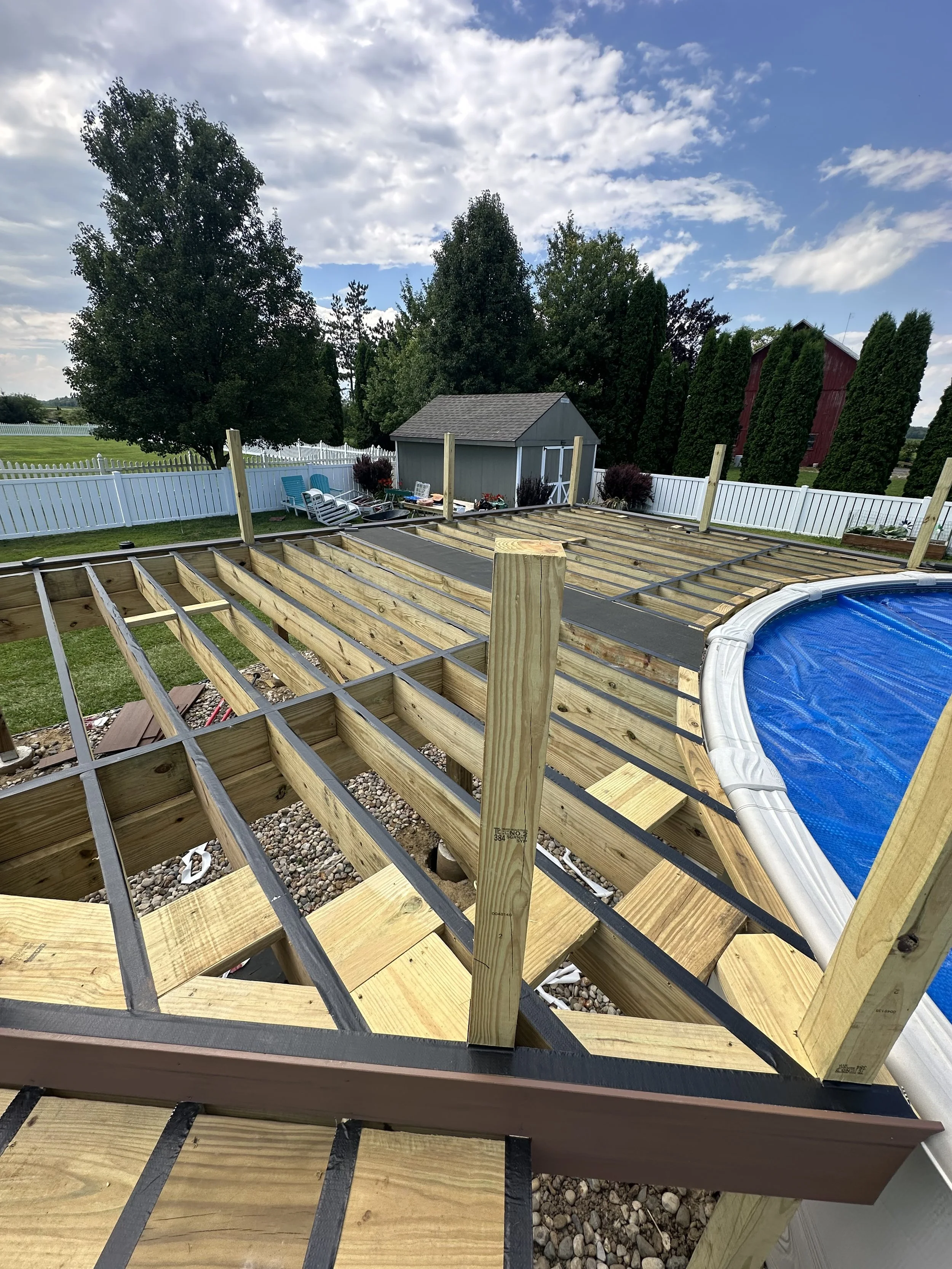 A backyard swimming pool under construction with wooden framework and a partially assembled deck area, surrounded by a white picket fence, trees, and a cloudy sky.