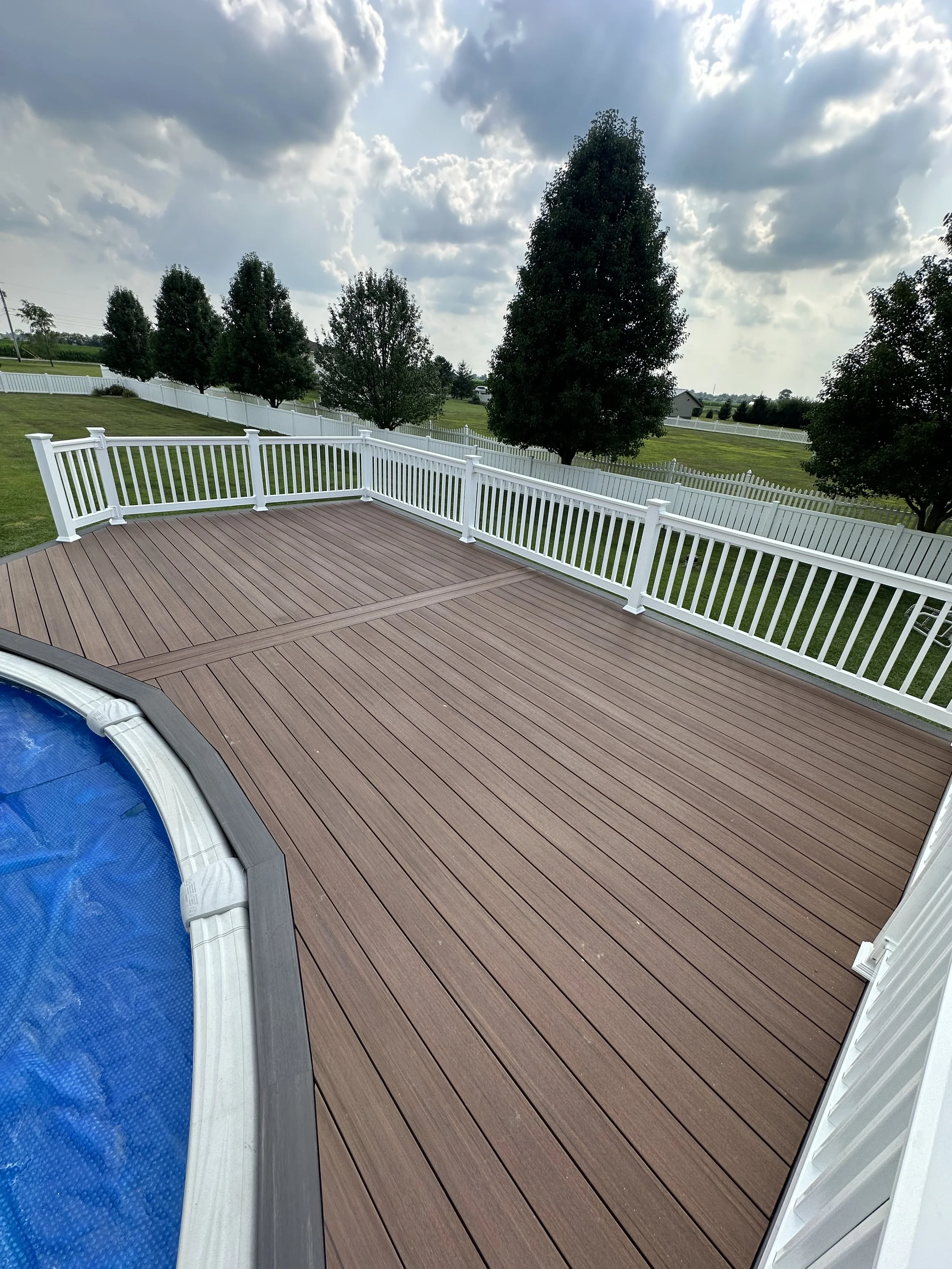 Empty backyard deck with a swimming pool and a white fence, surrounded by green trees and open sky with clouds.