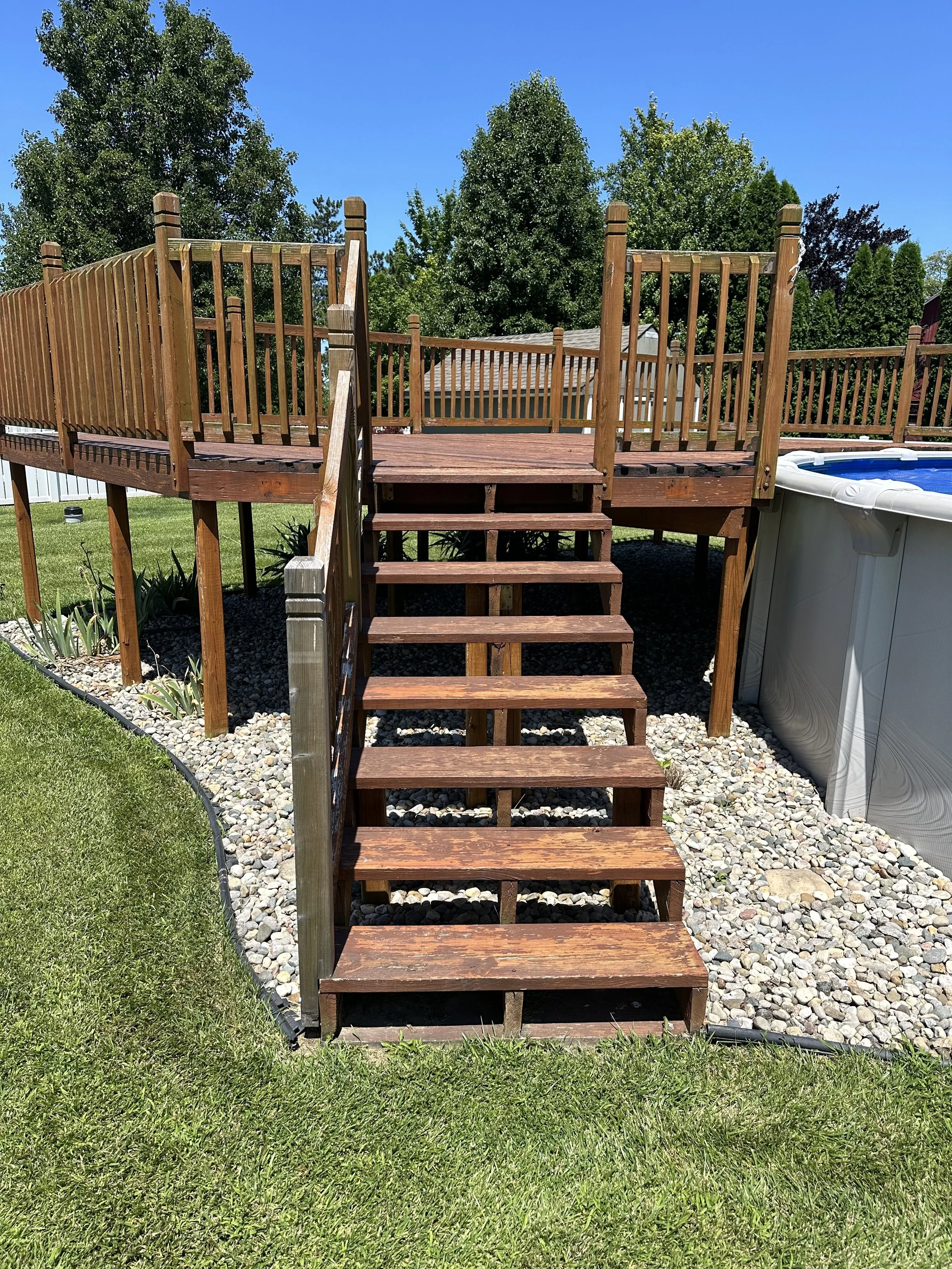 Backyard wooden deck with stairs leading down to a grassy lawn, surrounded by mulch and rocks, with trees and a blue sky in the background.