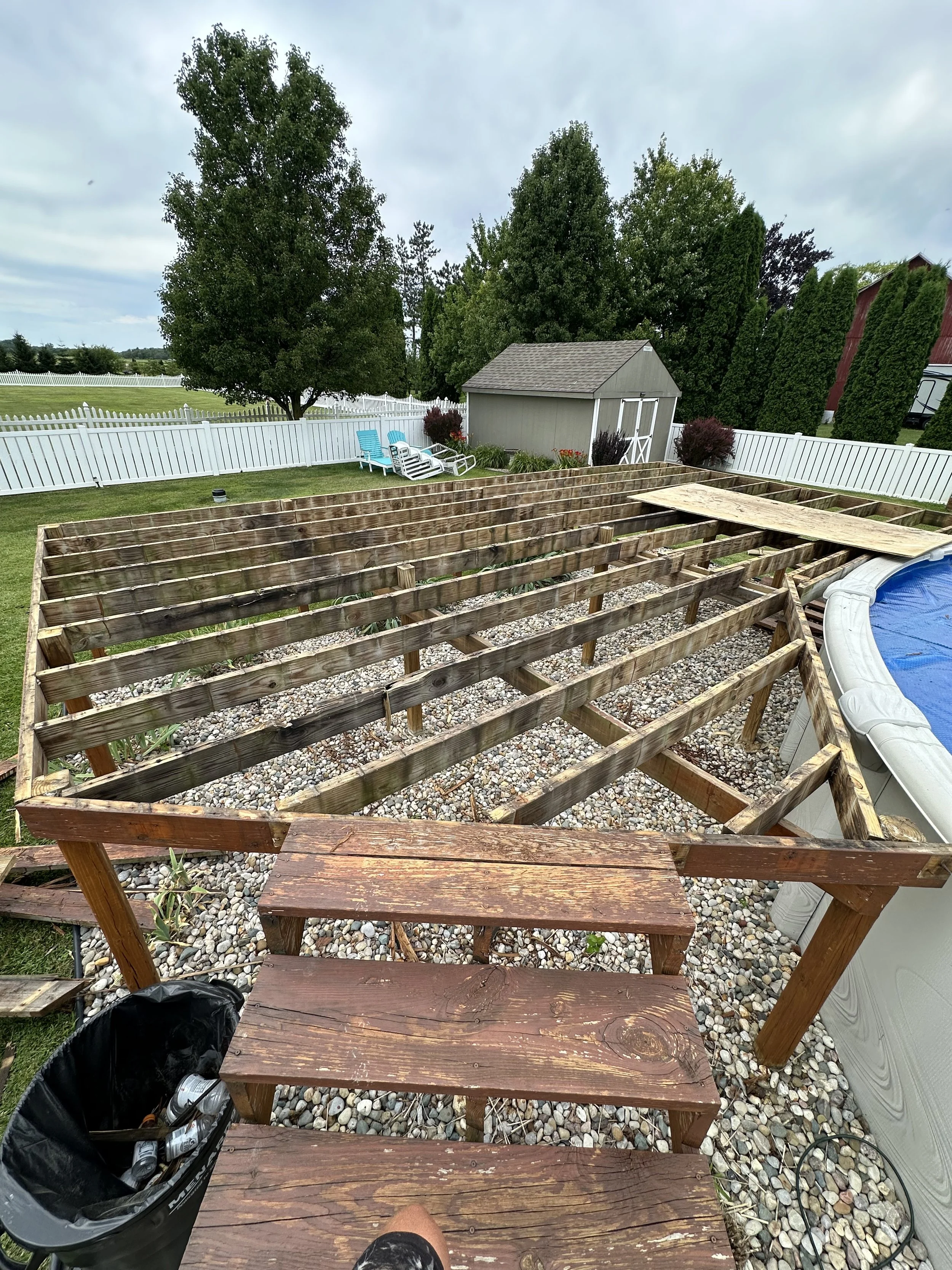 Backyard under construction with wooden deck framing, gravel ground, and a step ladder. In the background, there is a white fence, trees, a shed, and a small patio with blue chairs.