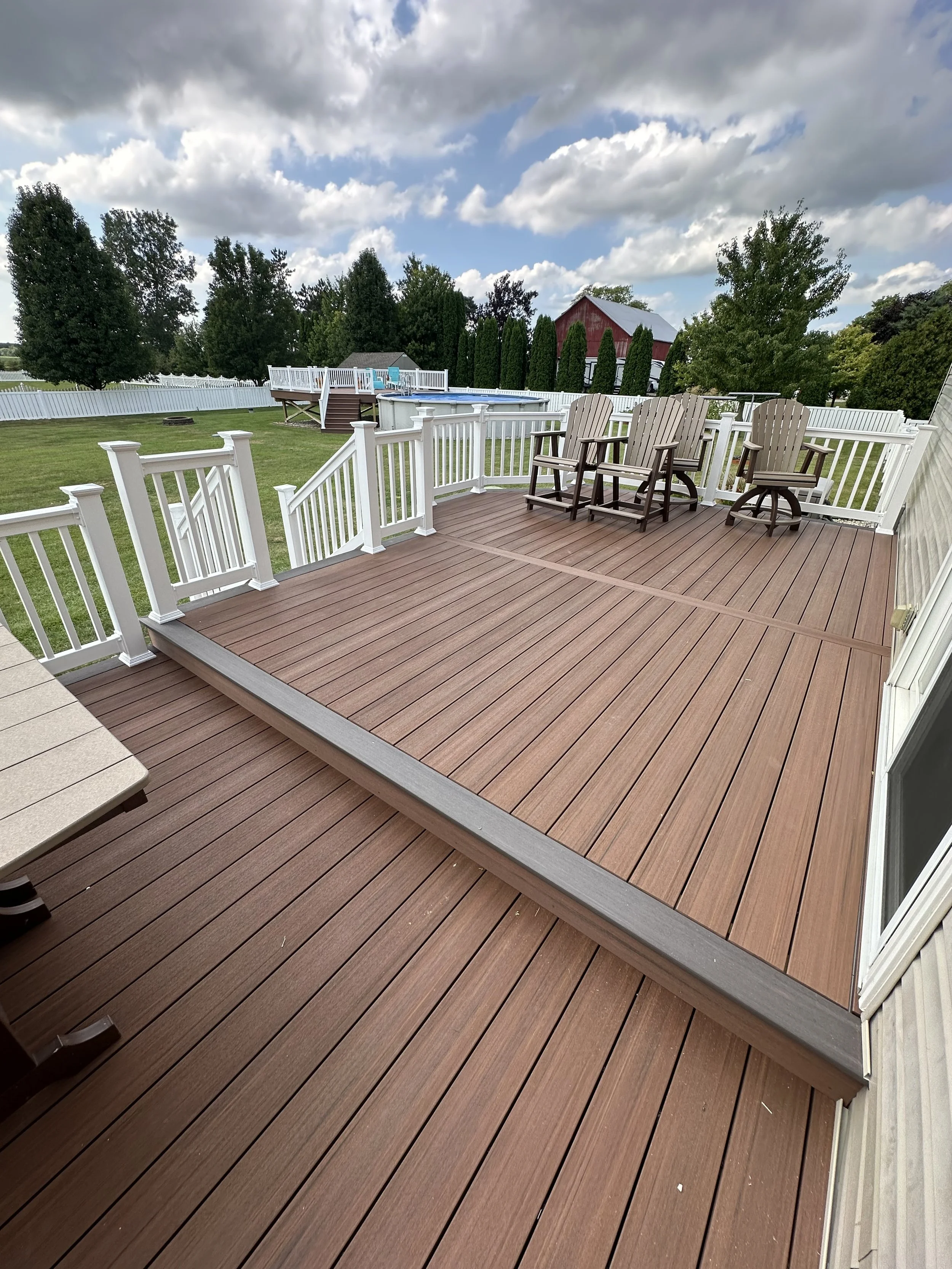 View of a spacious wooden deck with white railing, outdoor chairs, a grassy yard, a pool, and trees under a partly cloudy sky.