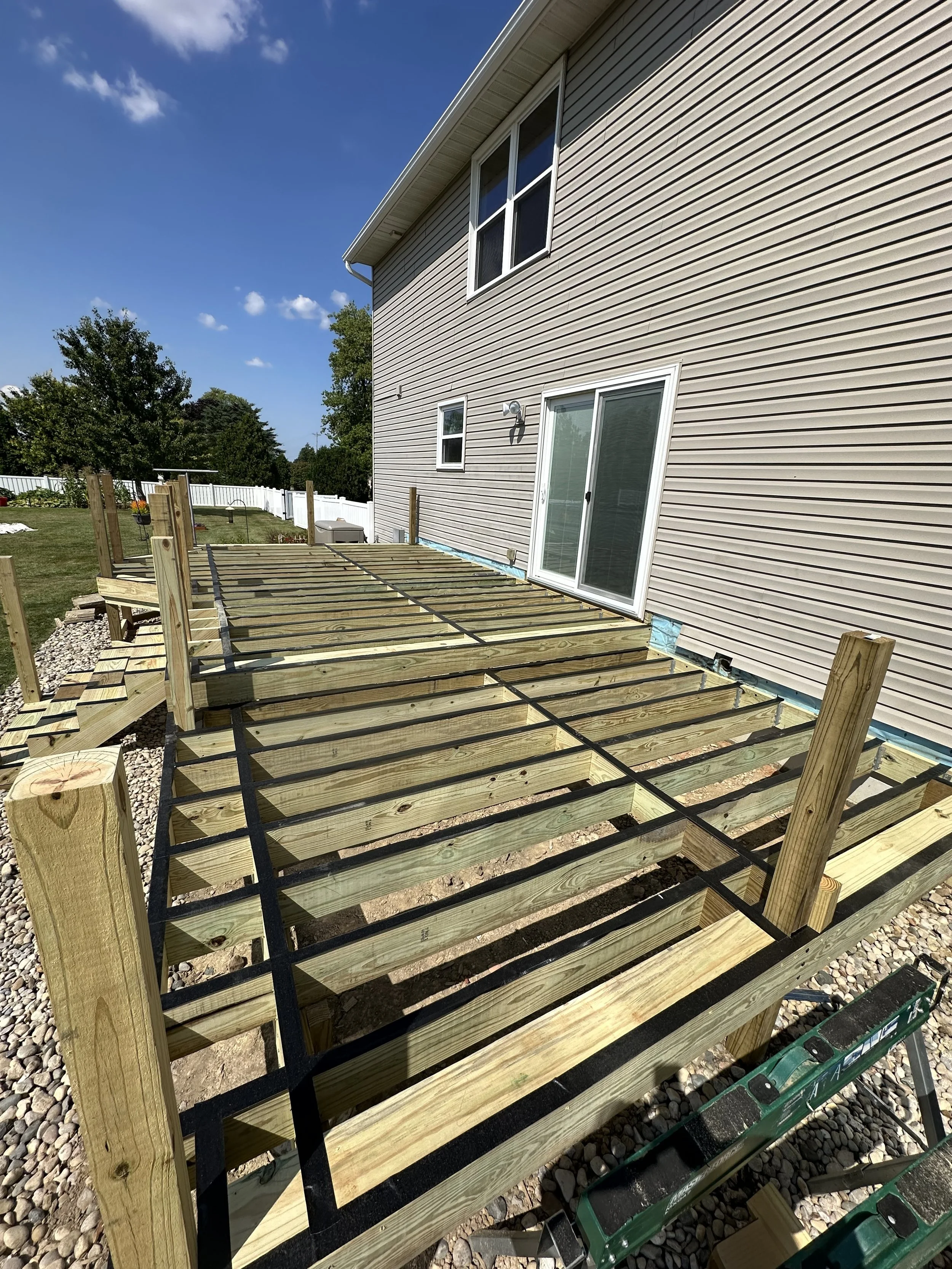 Deck construction in progress with wooden frame and black support beams attached to the house, under a clear blue sky.