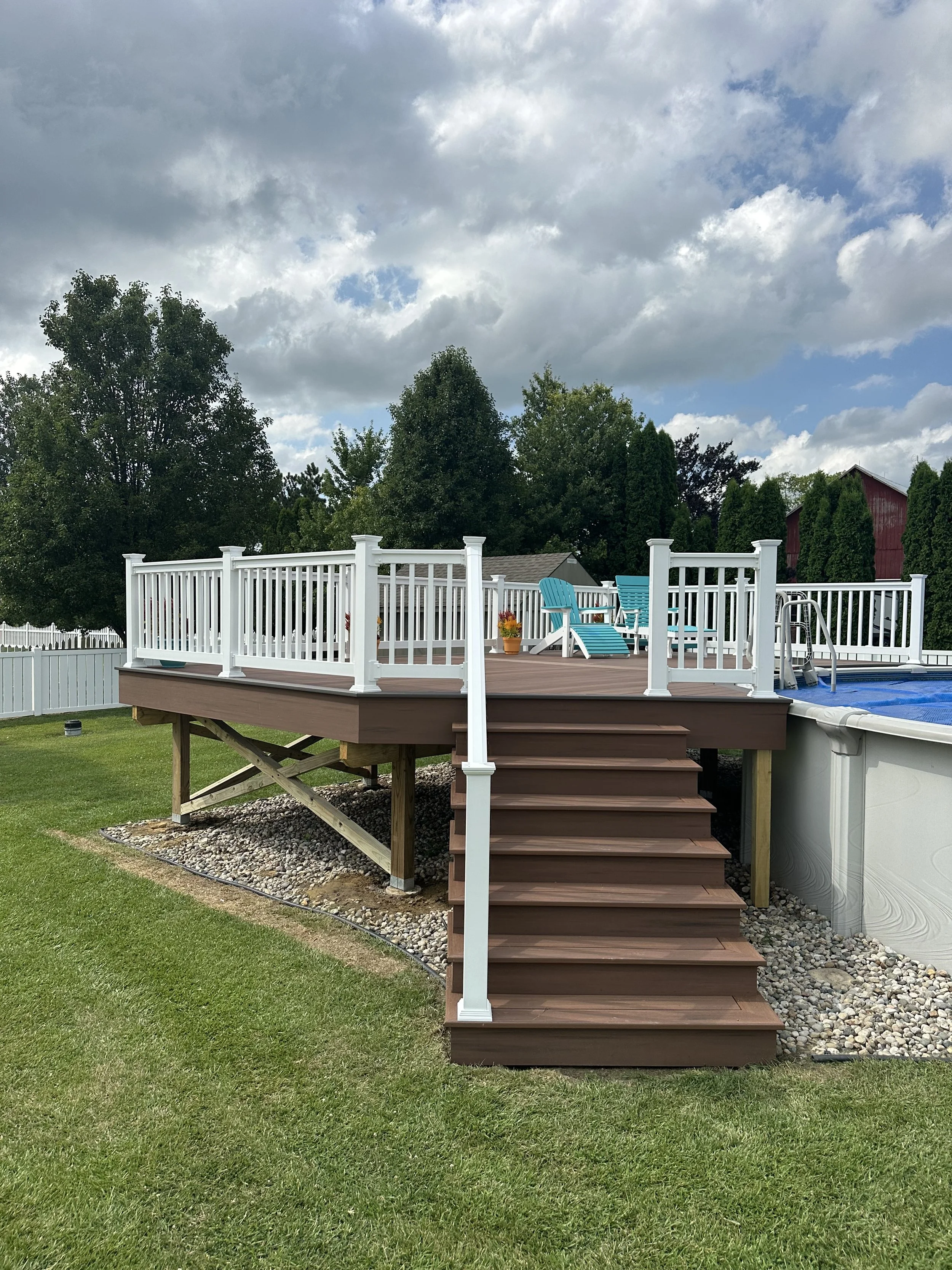 A backyard wooden deck with stairs leading up to it, surrounded by a white railing, with two blue chairs and potted plants on the deck, a swimming pool to the right, and trees in the background under a partly cloudy sky.