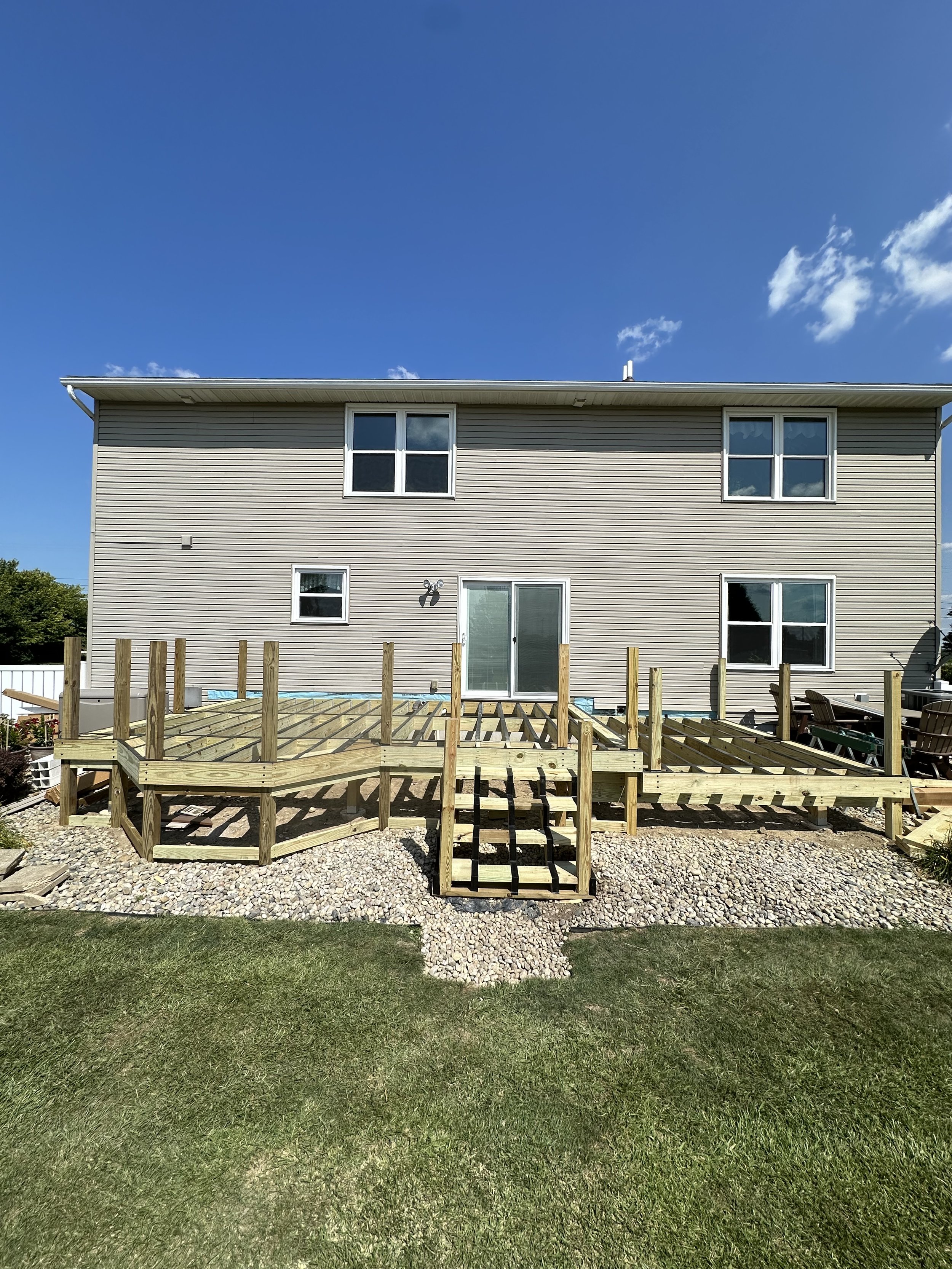 Backyard with a new wooden deck under construction in front of a beige house with four windows, sliding glass door, and a brick patio area, on a sunny day with blue sky and some clouds.