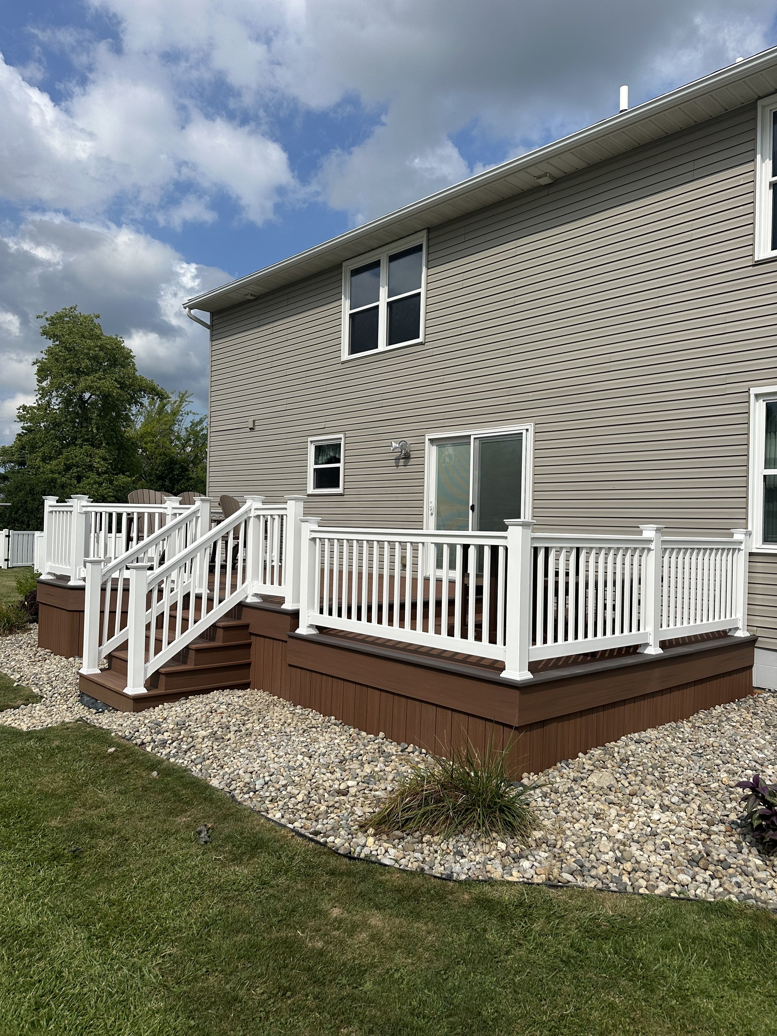 Backyard deck with white railing, brown steps, and surrounding gravel area, attached to a beige house with white-framed windows and sliding door, under partly cloudy sky.