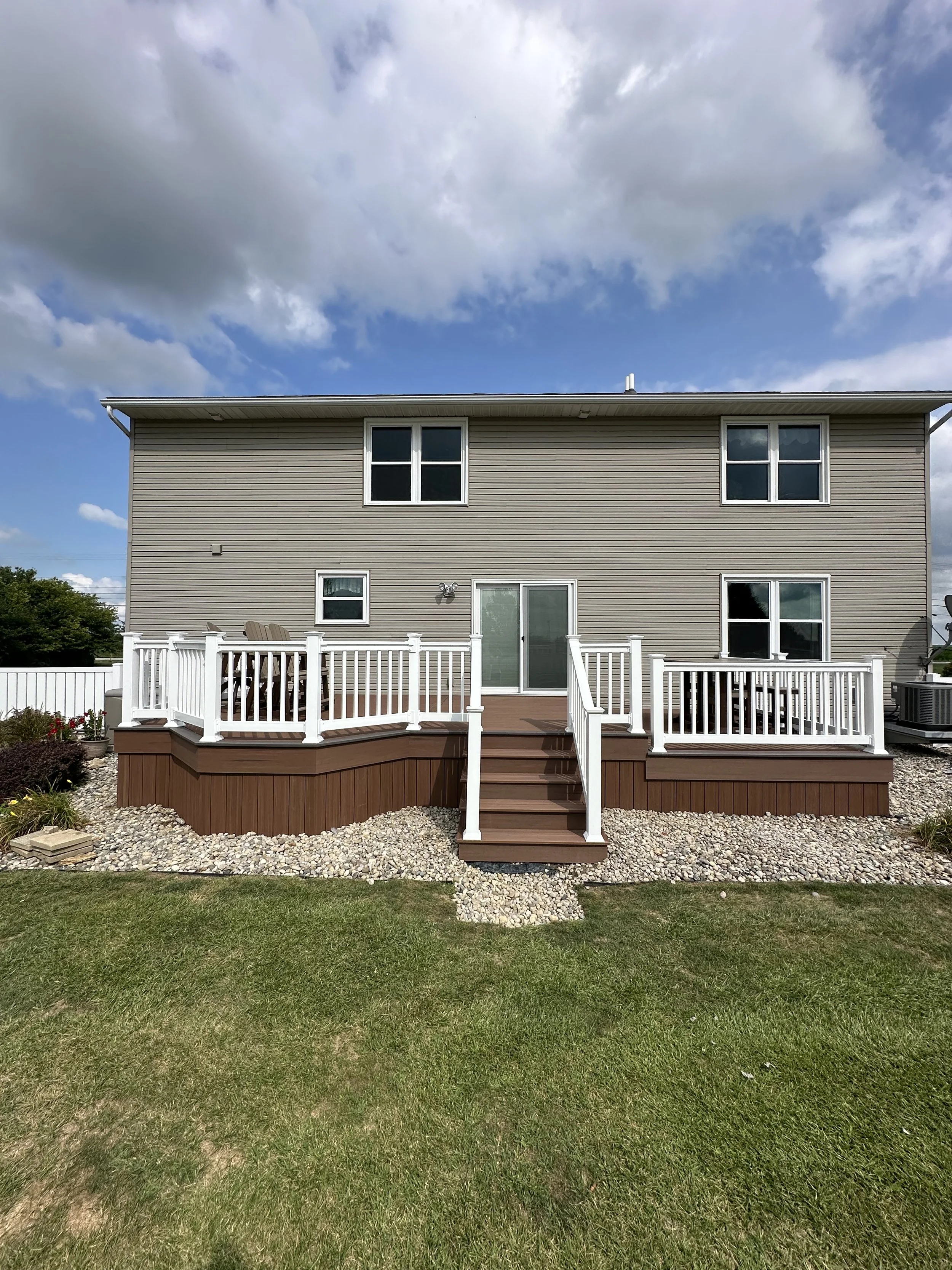 Backyard of a two-story house featuring a wooden deck with white railings, a sliding glass door, and four windows, with a grassy lawn and cloudy sky in the background.
