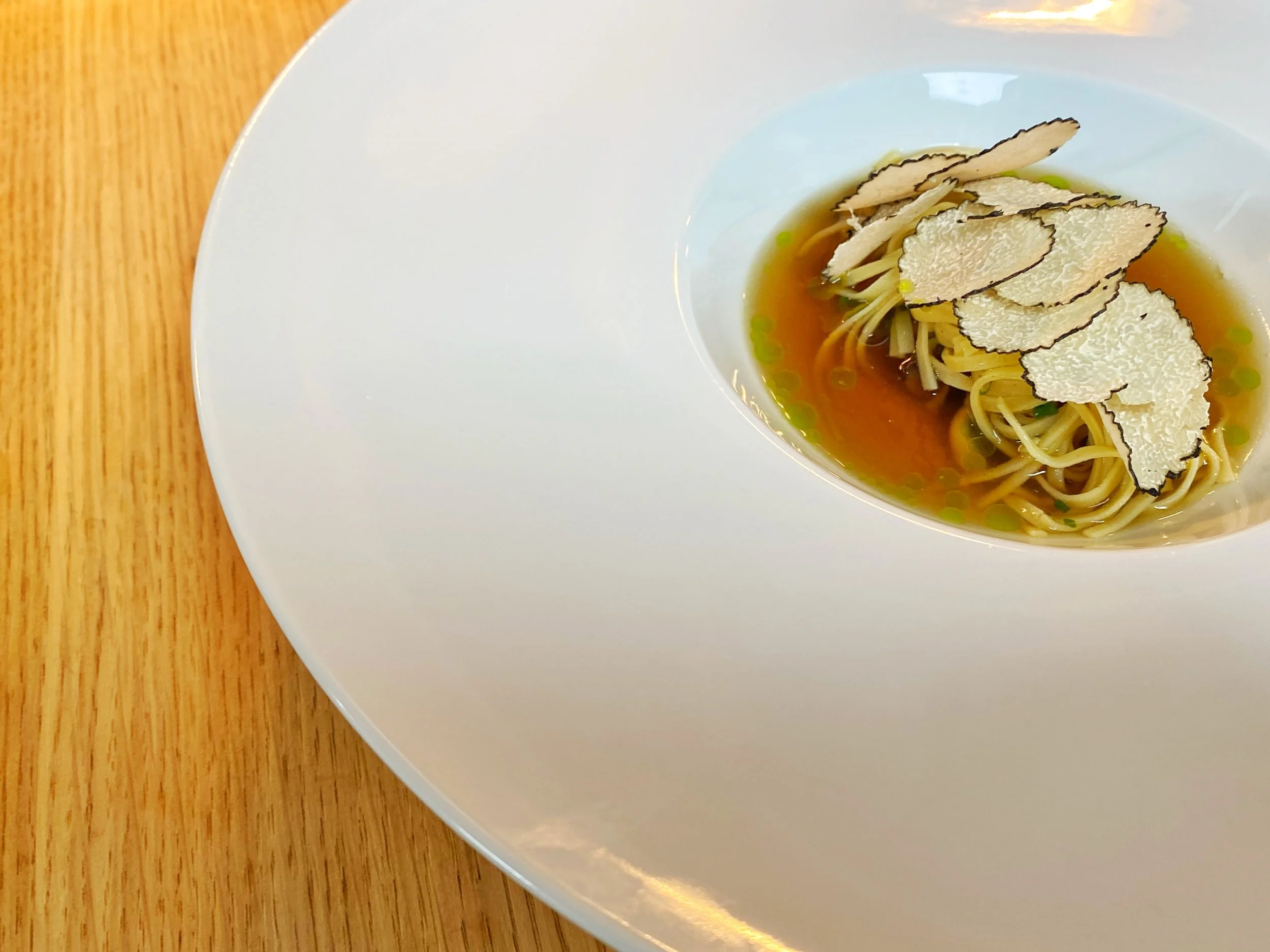 A white dish with a well of broth containing noodles and topped with sliced black truffles.