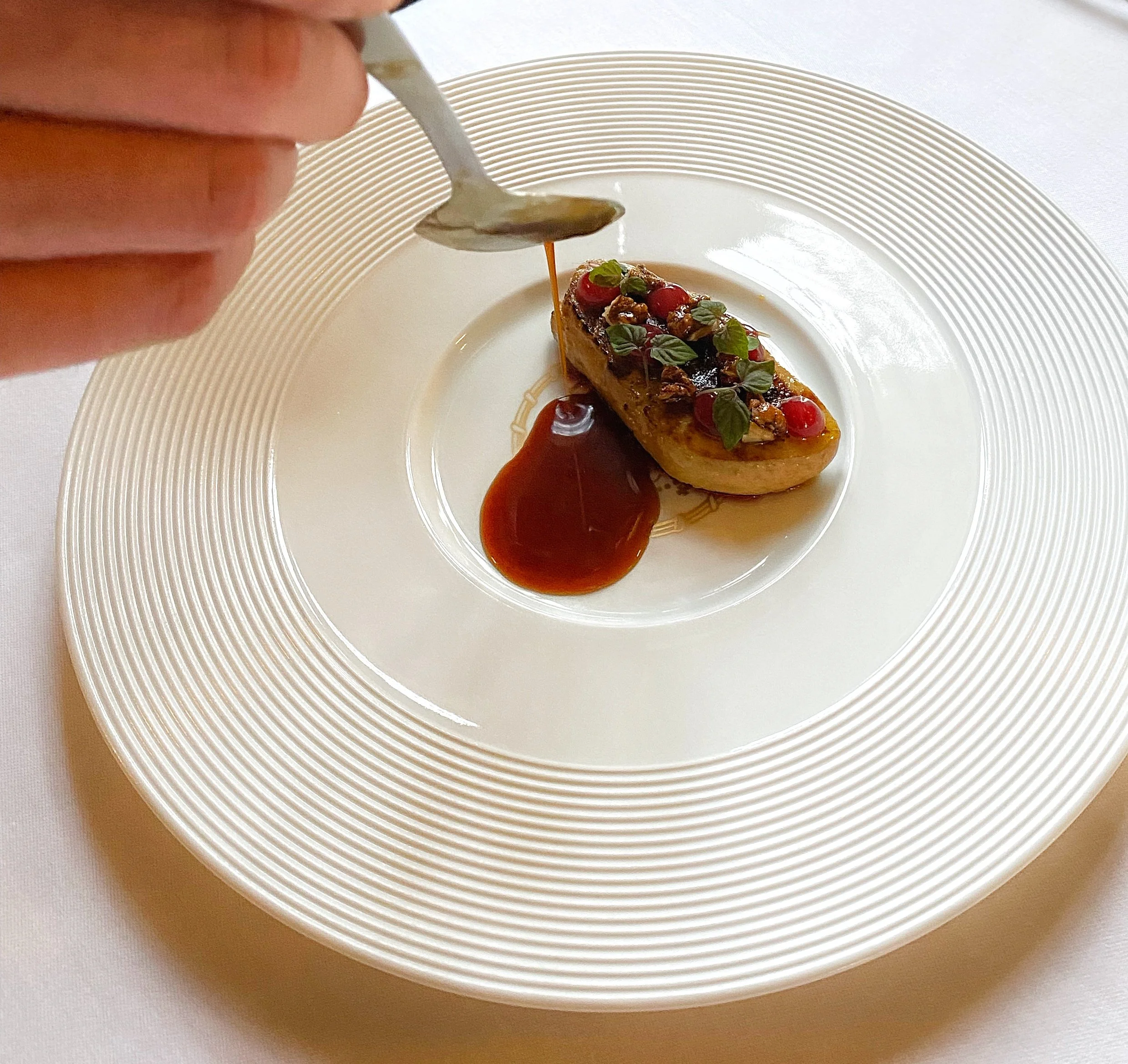 Dessert plate with a caramelized banana topped with berries, nuts, and mint leaves, with caramel sauce being drizzled over the top.