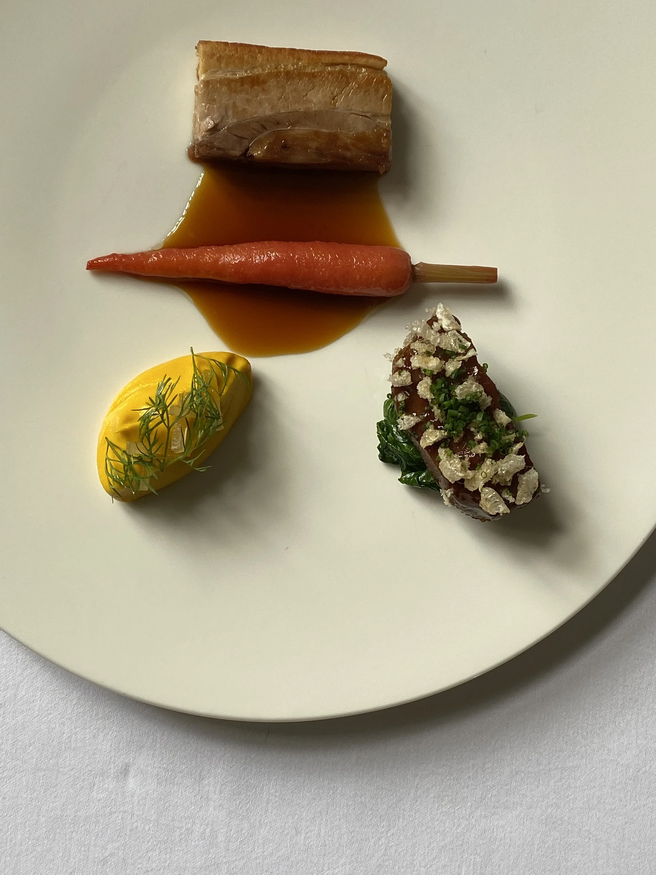 Gourmet dish with a piece of seared fish, a carrot with sauce, a dollop of yellow puree topped with herbs, and a small portion of meat topped with white garnish on a white plate.