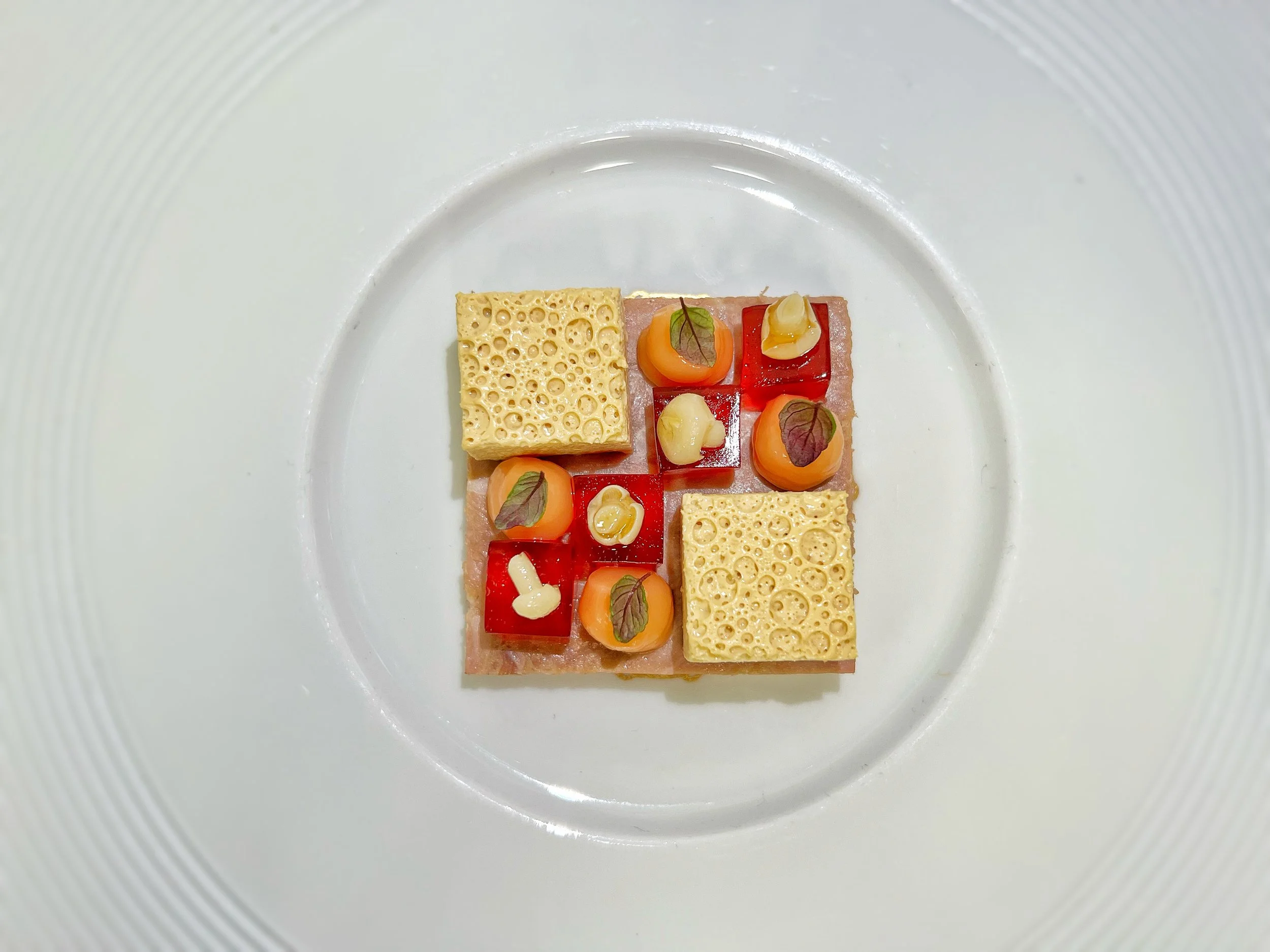 A modern, artfully plated dish featuring a square base of pinkish meat, topped with small orange and red vegetables, garnished with small leaves, and accented with two pieces of cheese and small dollops of a creamy substance on a white plate.