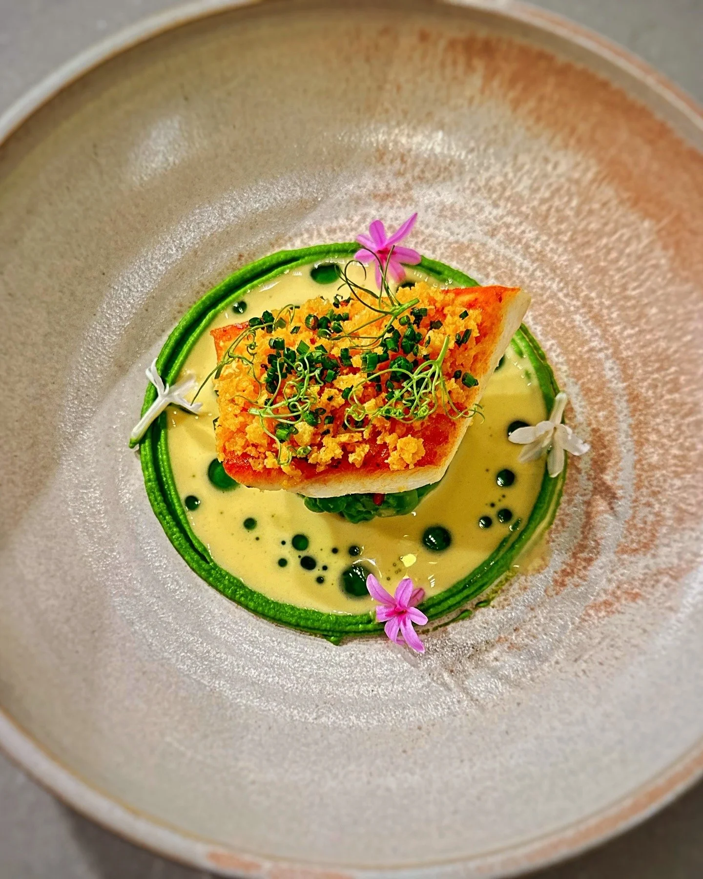 Elegant dish with a piece of grilled fish on green vegetables, topped with microgreens and edible flowers, served on a creamy sauce with black droplets, garnished with a green spiral and pink narcissus flowers.