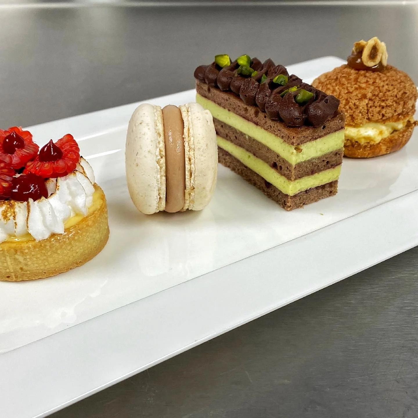 Four assorted desserts on a white rectangular platter, including a fruit-topped tart, a macaroon, a layered cake with green filling and chocolate topping, and a cream puff with a small piece of hazelnut on top.