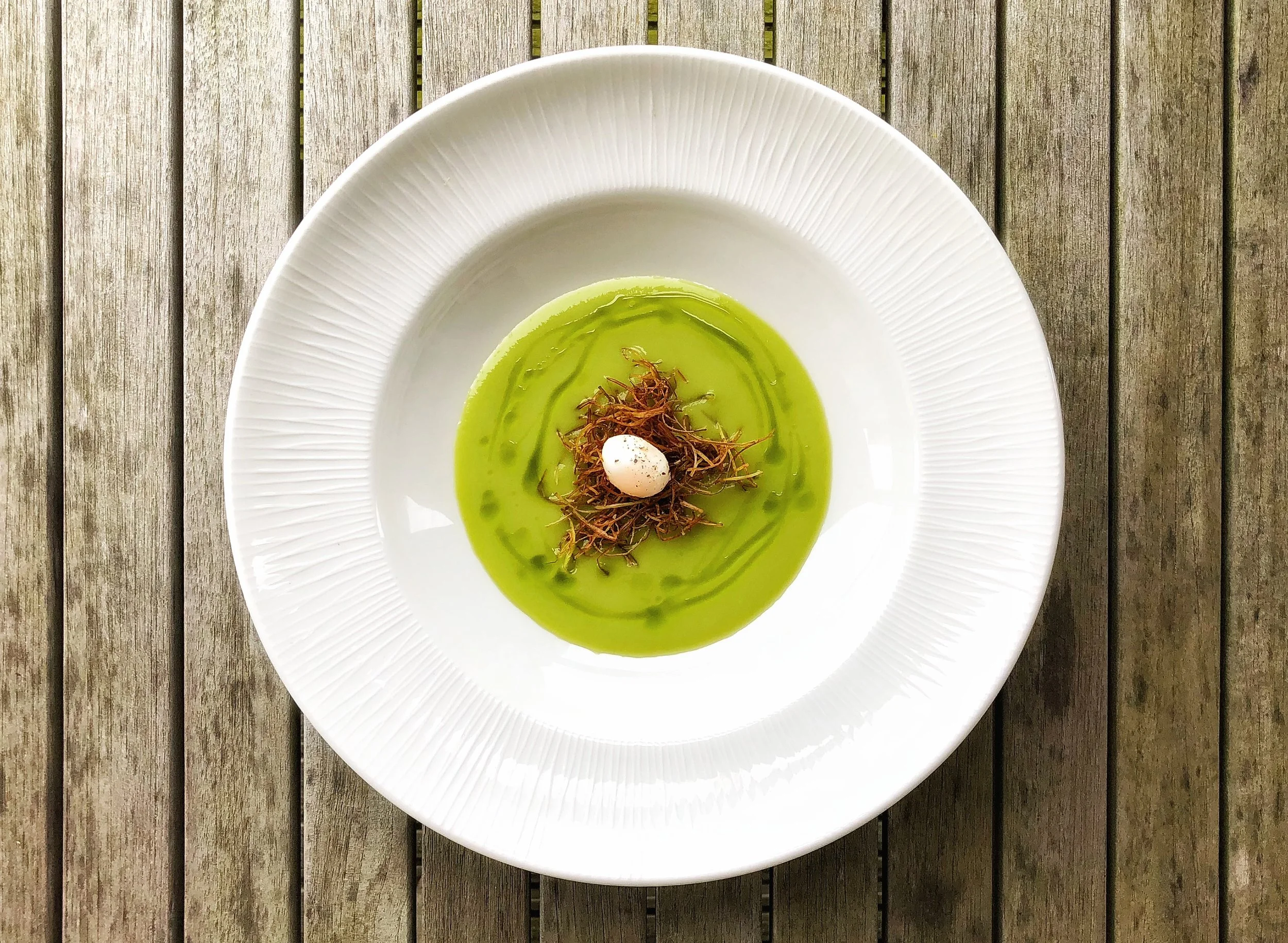 A white plate with a green soup garnished with crispy herbs and a small white quail egg on top, placed on a wooden surface.