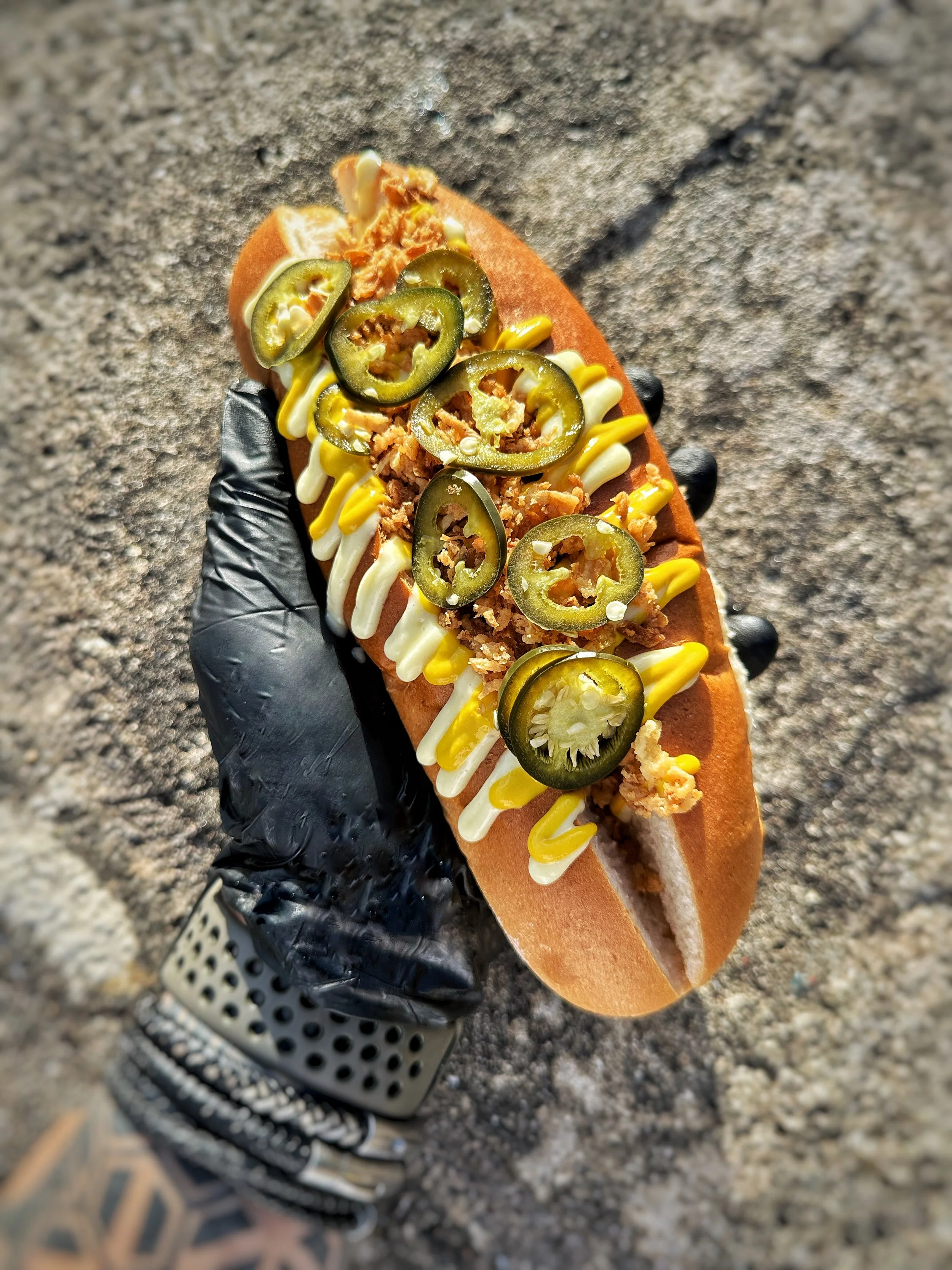 Hand wearing a black glove holding a hot dog in a bun topped with yellow mustard, white sauce, crushed chips, and sliced jalapeño peppers, on a sandy ground.
