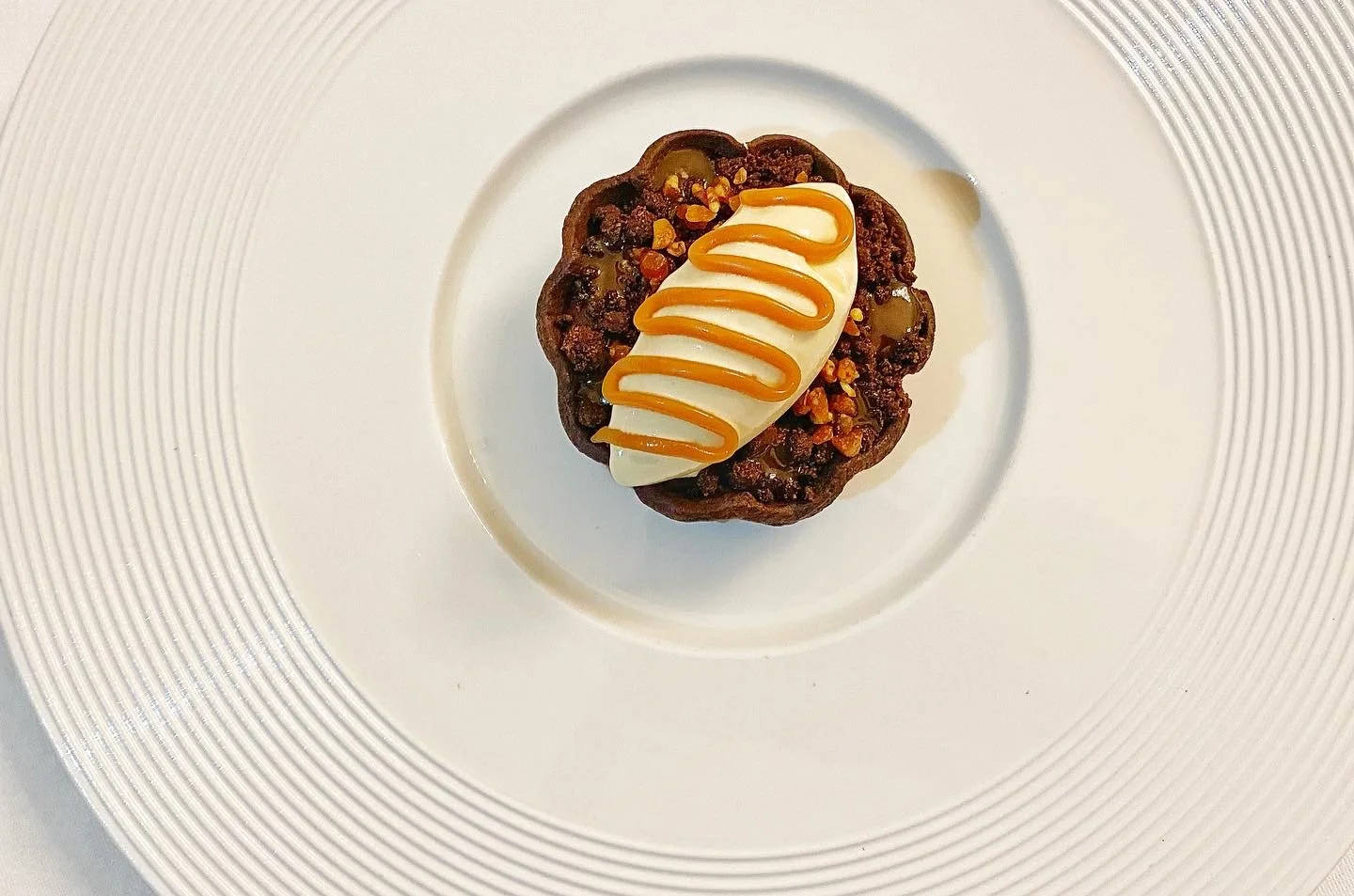 Dessert with a chocolate tart topped with a dollop of cream or mousse, drizzled caramel sauce, served on a white plate.