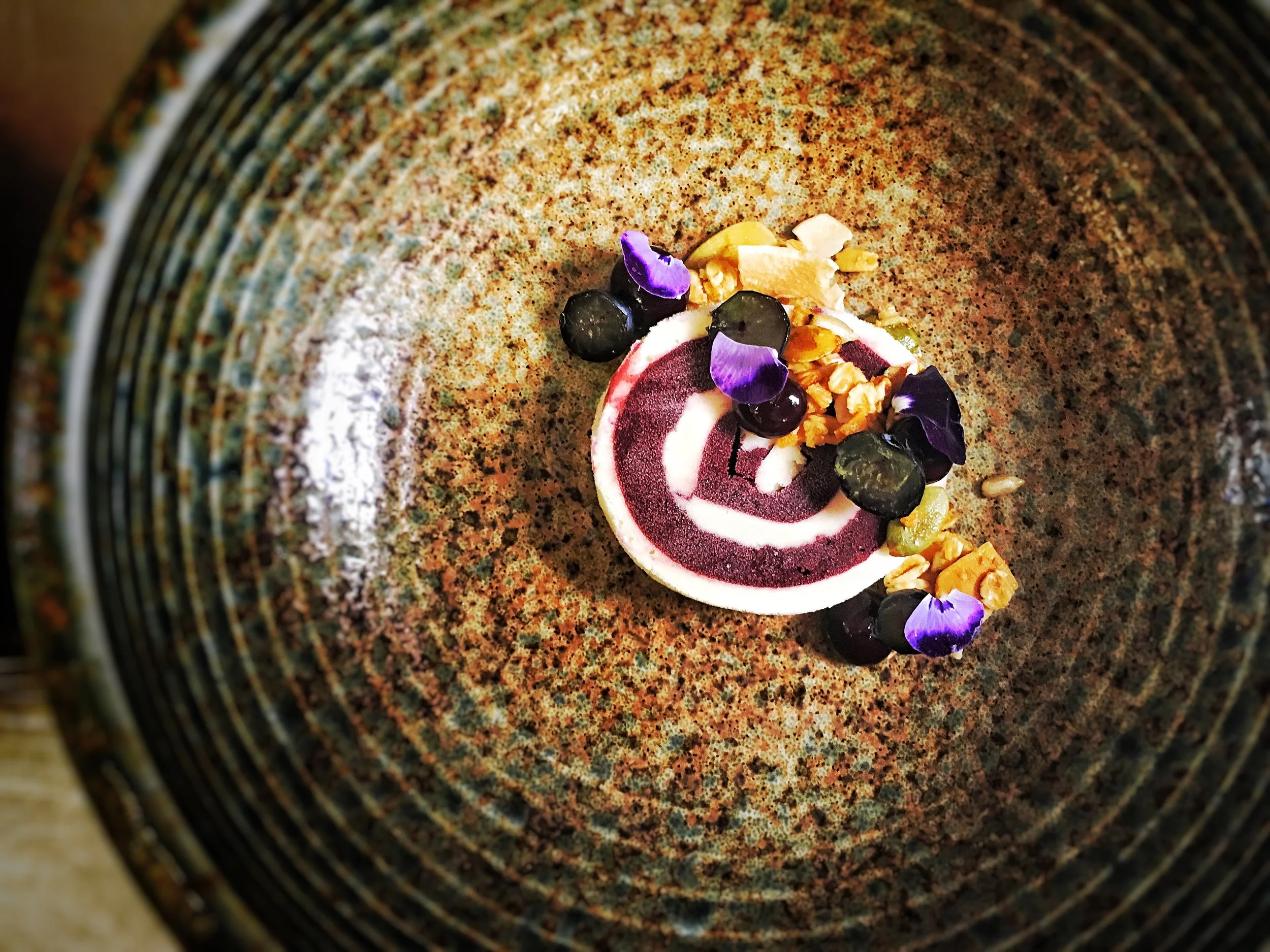 A swirl of white and purple mousse on a textured brown plate, garnished with blueberries, granola, and purple edible flower petals.