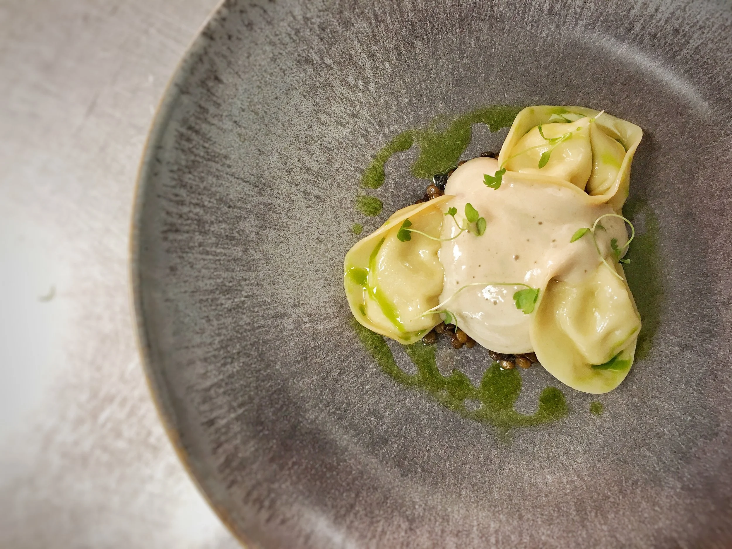A plated gourmet dish featuring stuffed pasta with a creamy sauce, garnished with microgreens, served on a dark textured plate.