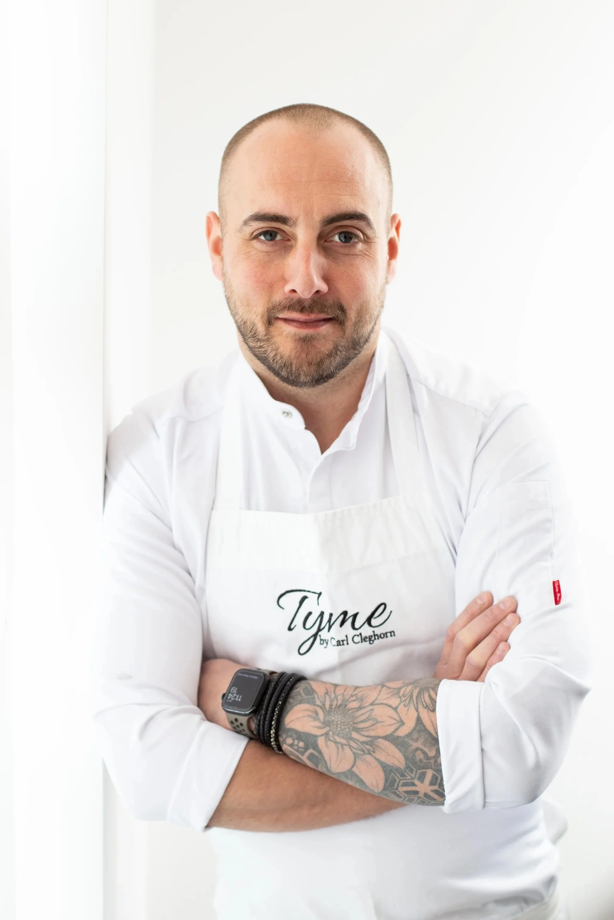 welsh chef Carl Cleghorn,with a beard and tattoos on his left arm, wearing a white chef's coat and apron, standing with arms crossed, in a bright white setting. private chef