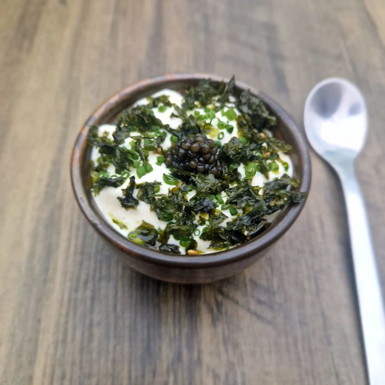A bowl of creamy yogurt topped with chopped green onions, salted seaweed, and black caviar, with a spoon placed beside it on a wooden table.