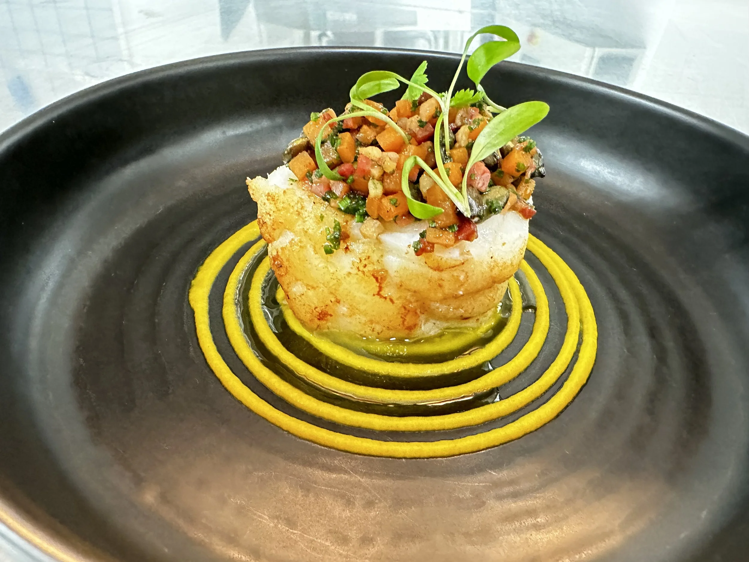 A plate with a piece of bbq monkfish topped with pickled pumpkin, pumpkin seeds and coriander, garnished with microgreens, presented on a black plate with pumpkin puree rings underneath.