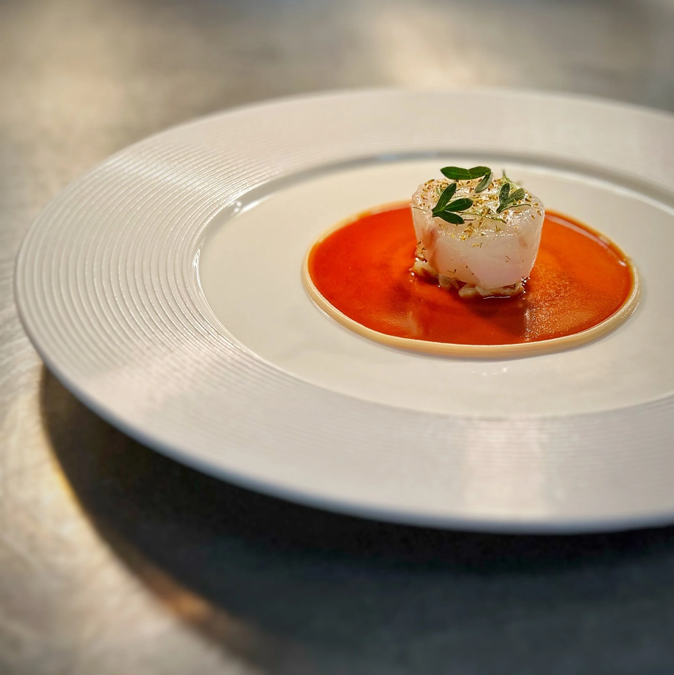 A gourmet dish featuring a seared scallop garnished with greens, served on a bed of orange sauce on a white, textured plate.