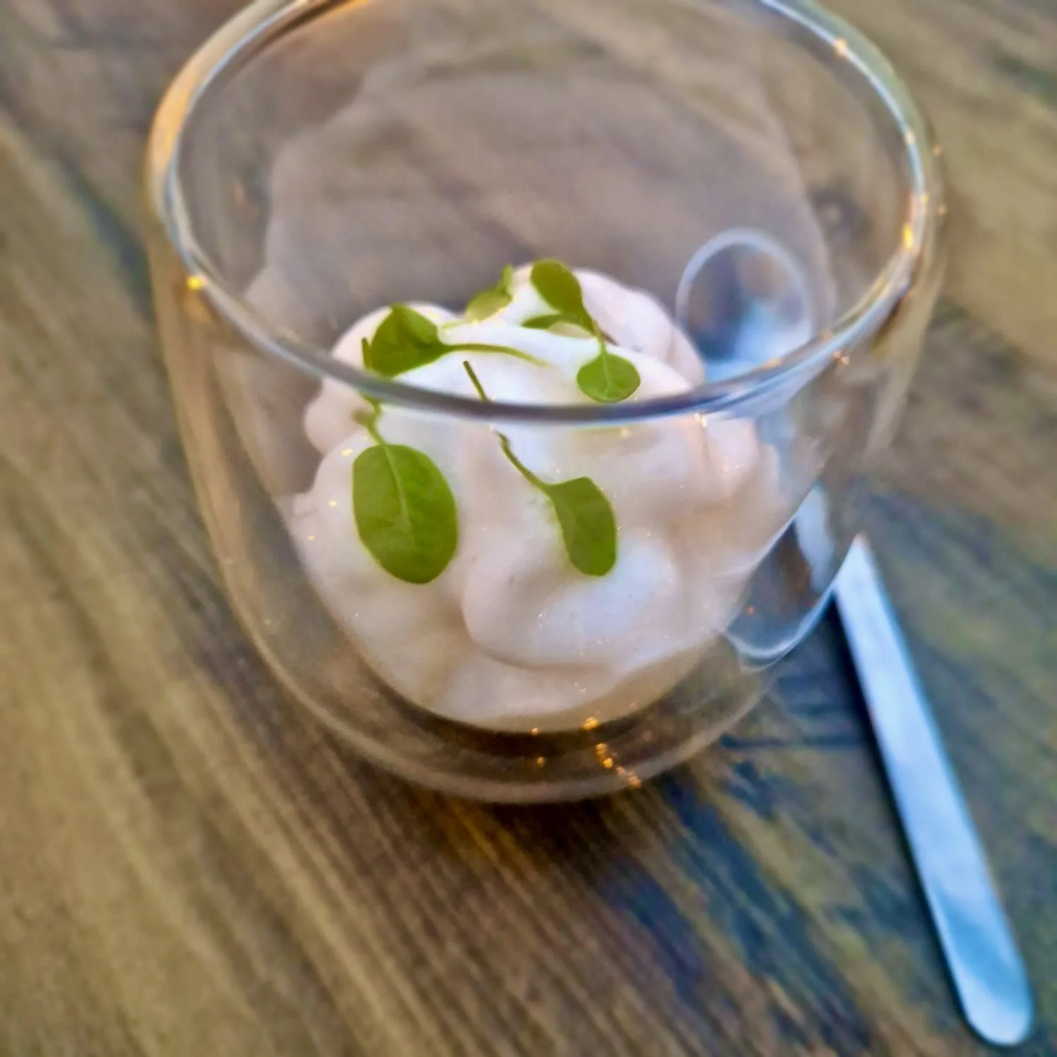 A clear glass cup with whipped cream and green herbs on top, placed on a wooden table with a fork beside it.