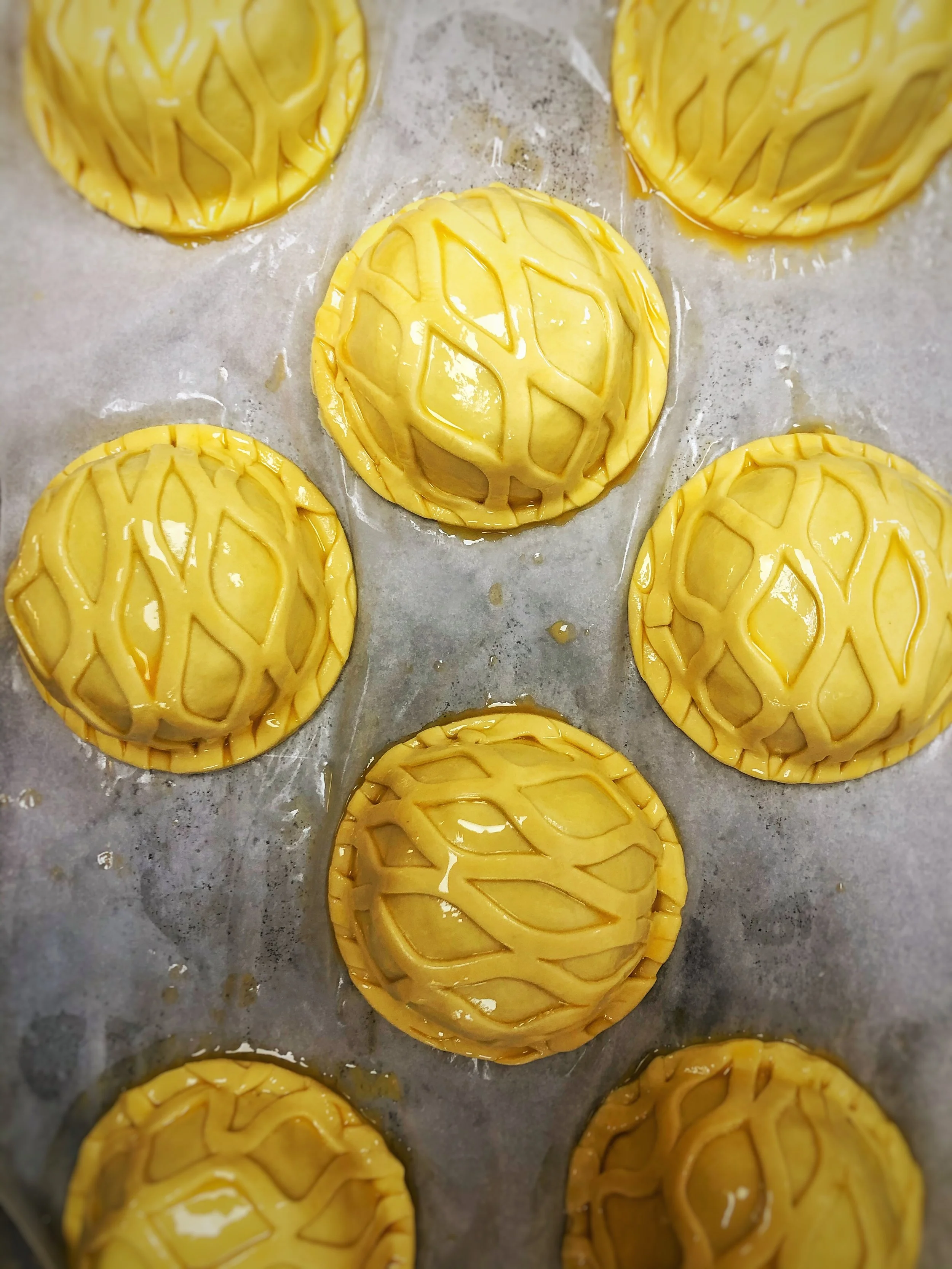 Unbaked mini cheeseburgers with decorative lattice tops on a baking sheet lined with parchment paper.